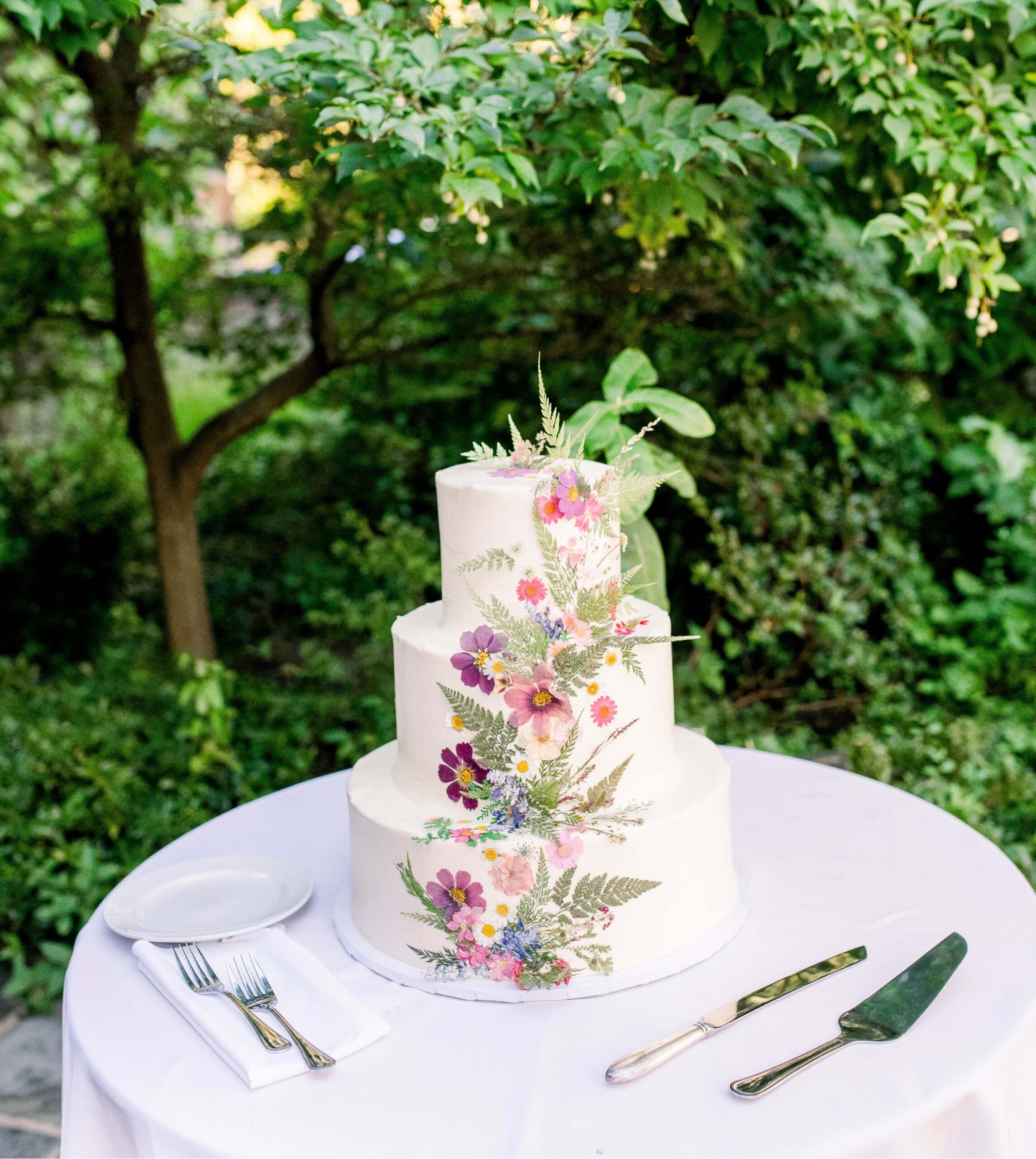 Three-tiered white cake with colorful floral decorations sits on a white table. Plates, a knife, and a fork are arranged nearby. A lush green background surrounds the setup.