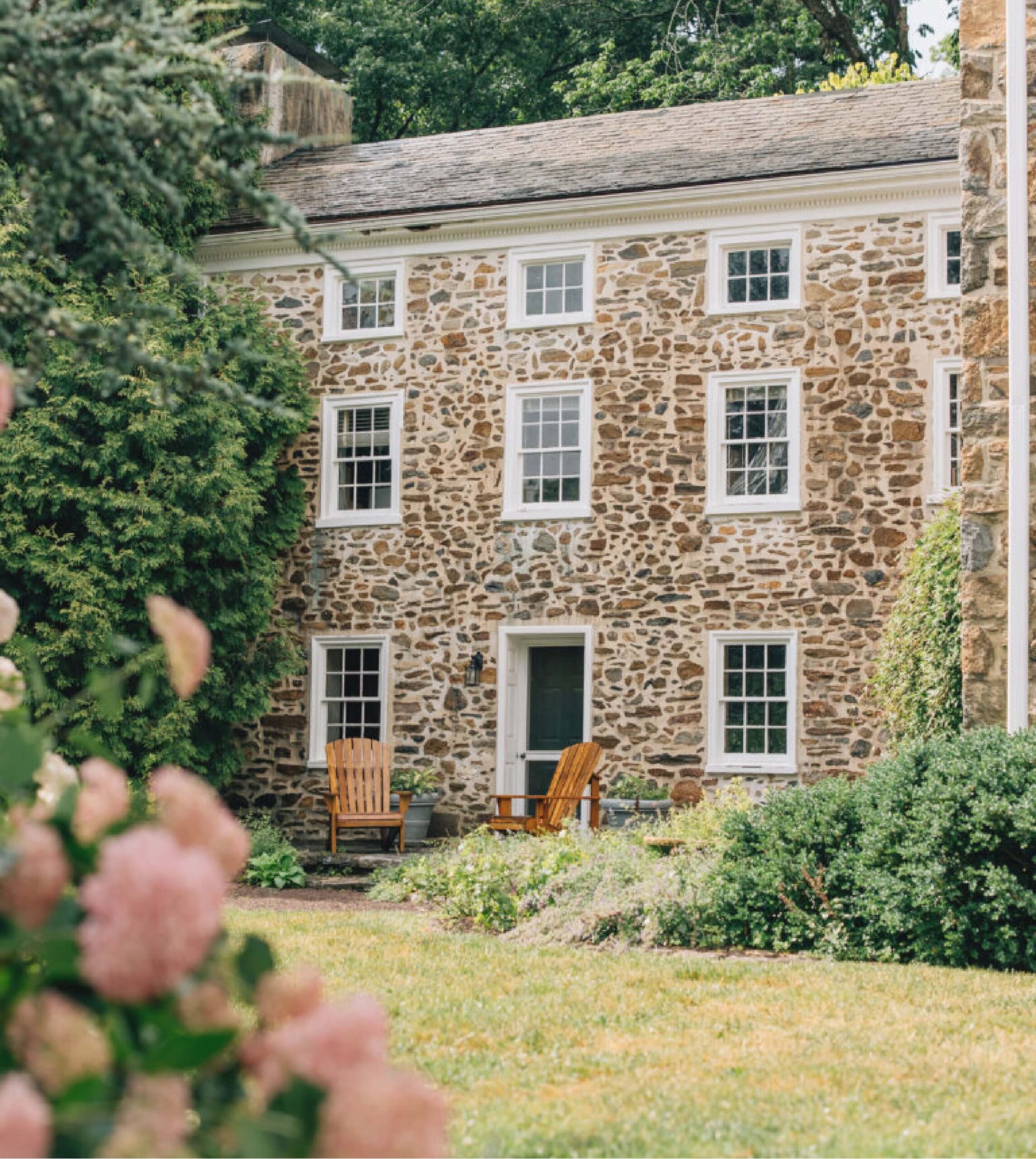 A stone house with multiple windows and a wooden door, surrounded by greenery and pink flowers. Two wooden chairs are placed near the entrance.