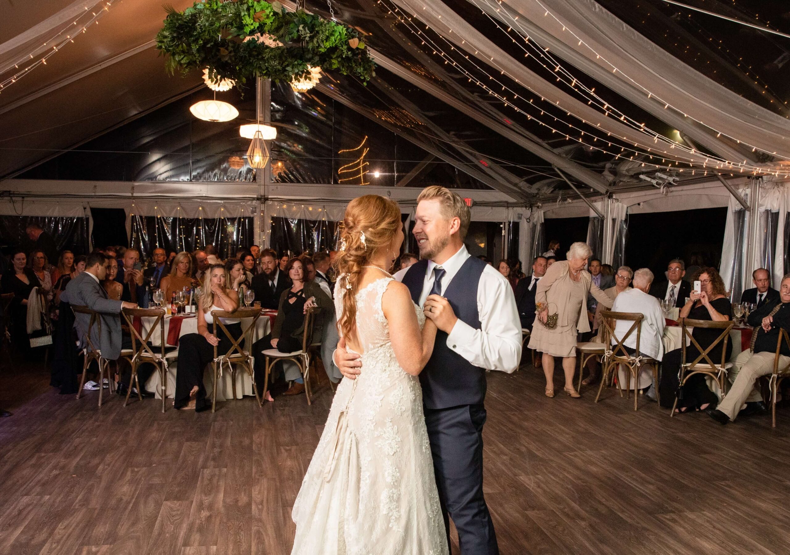 A bride and groom share a first dance under a tent decorated with string lights, surrounded by seated guests.