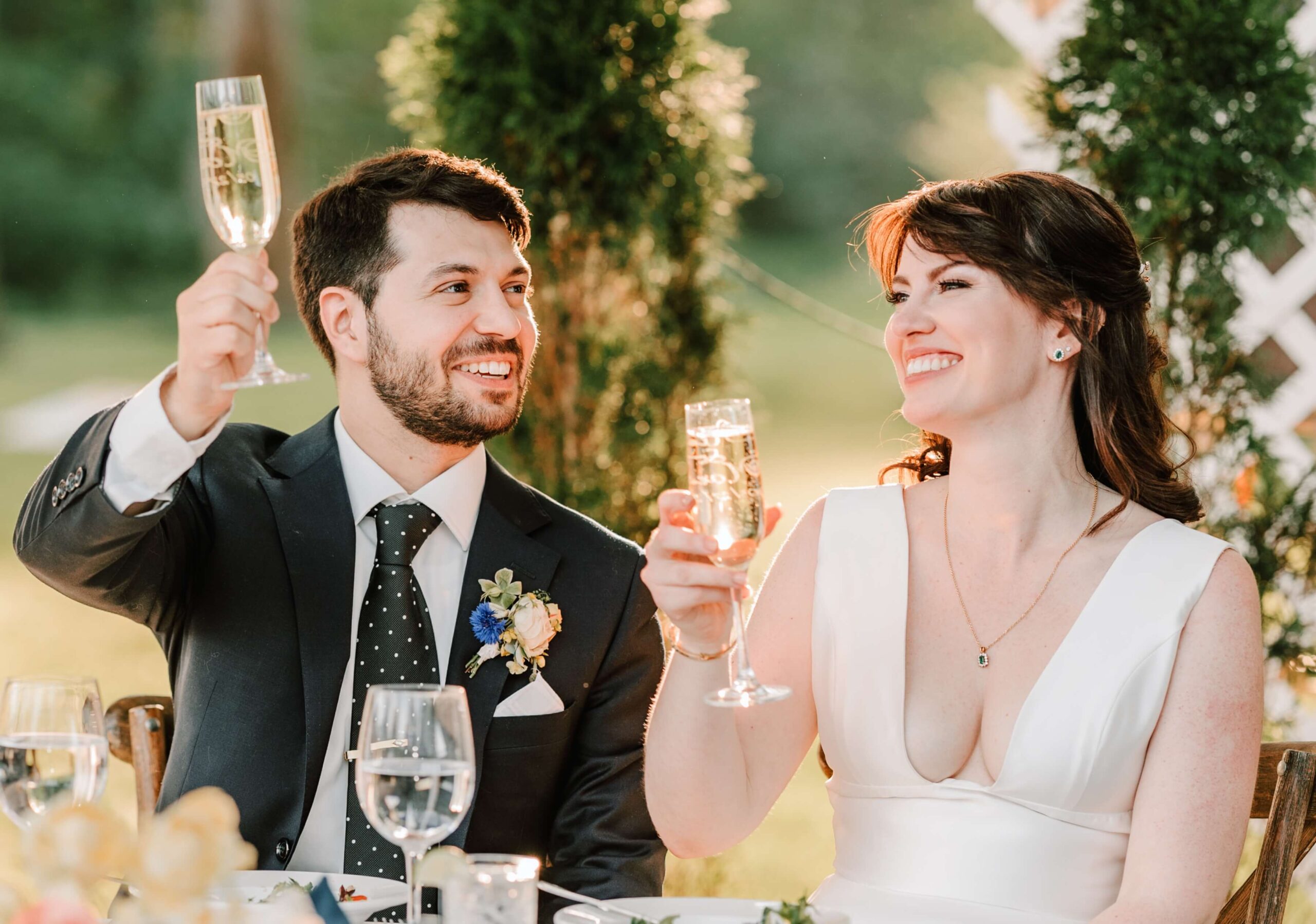 A couple sits at a table outdoors, holding champagne glasses in a toast. The man wears a suit, and the woman is in a white dress. They are smiling at each other.