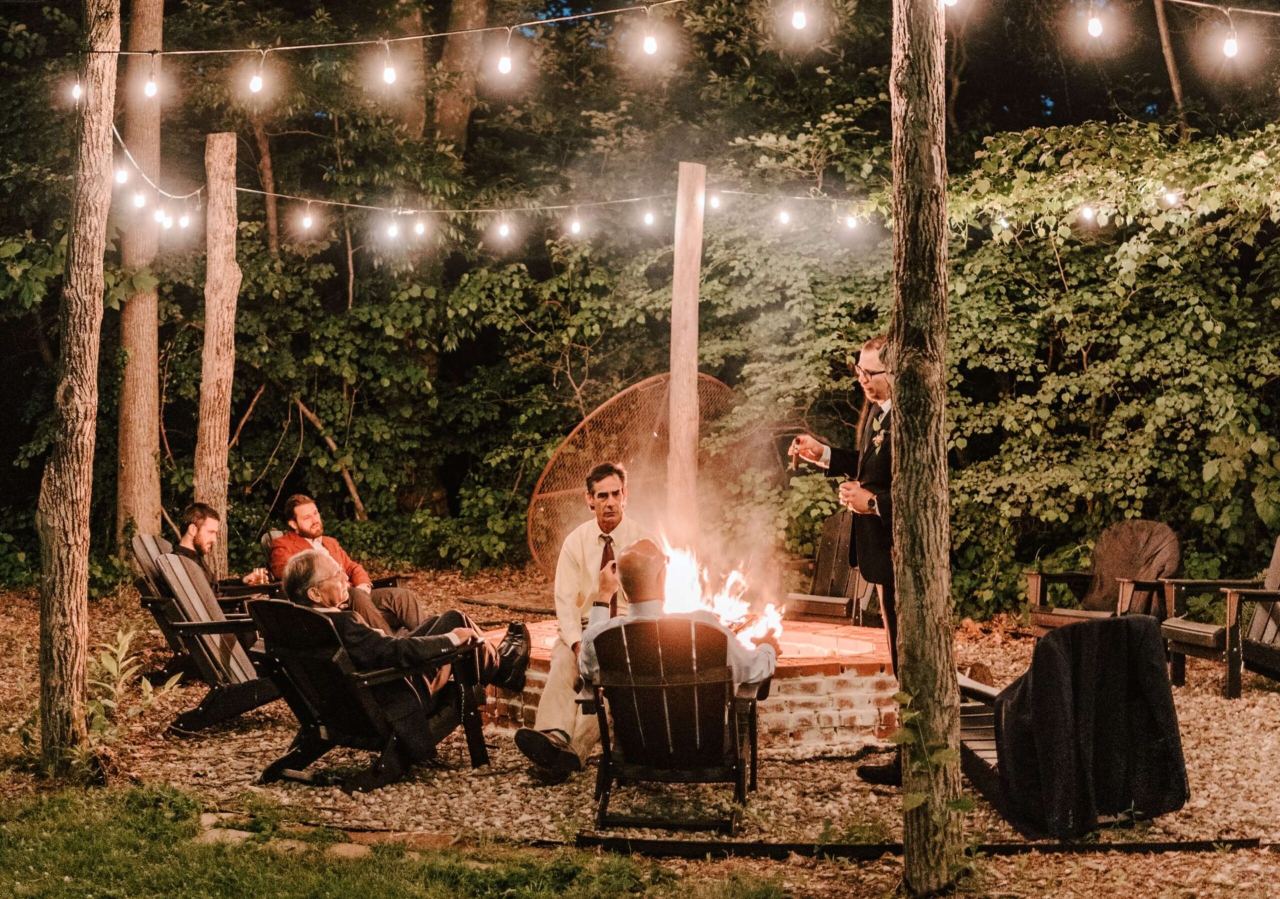 People sitting around a fire pit at night under string lights in a wooded area.
