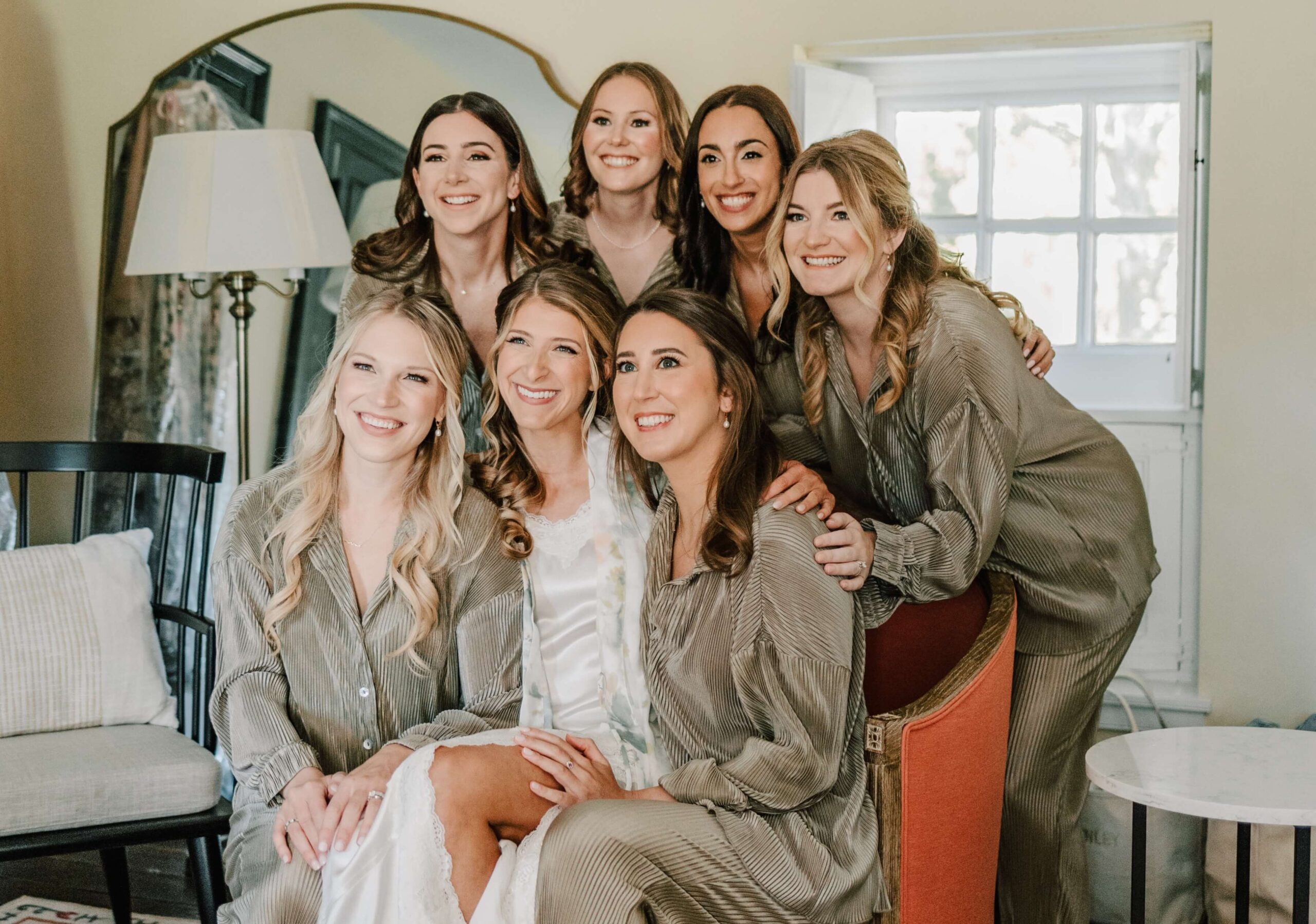 Seven women in matching attire pose together indoors, smiling. Some are seated while others stand behind them. A lamp and a mirror are visible in the background.