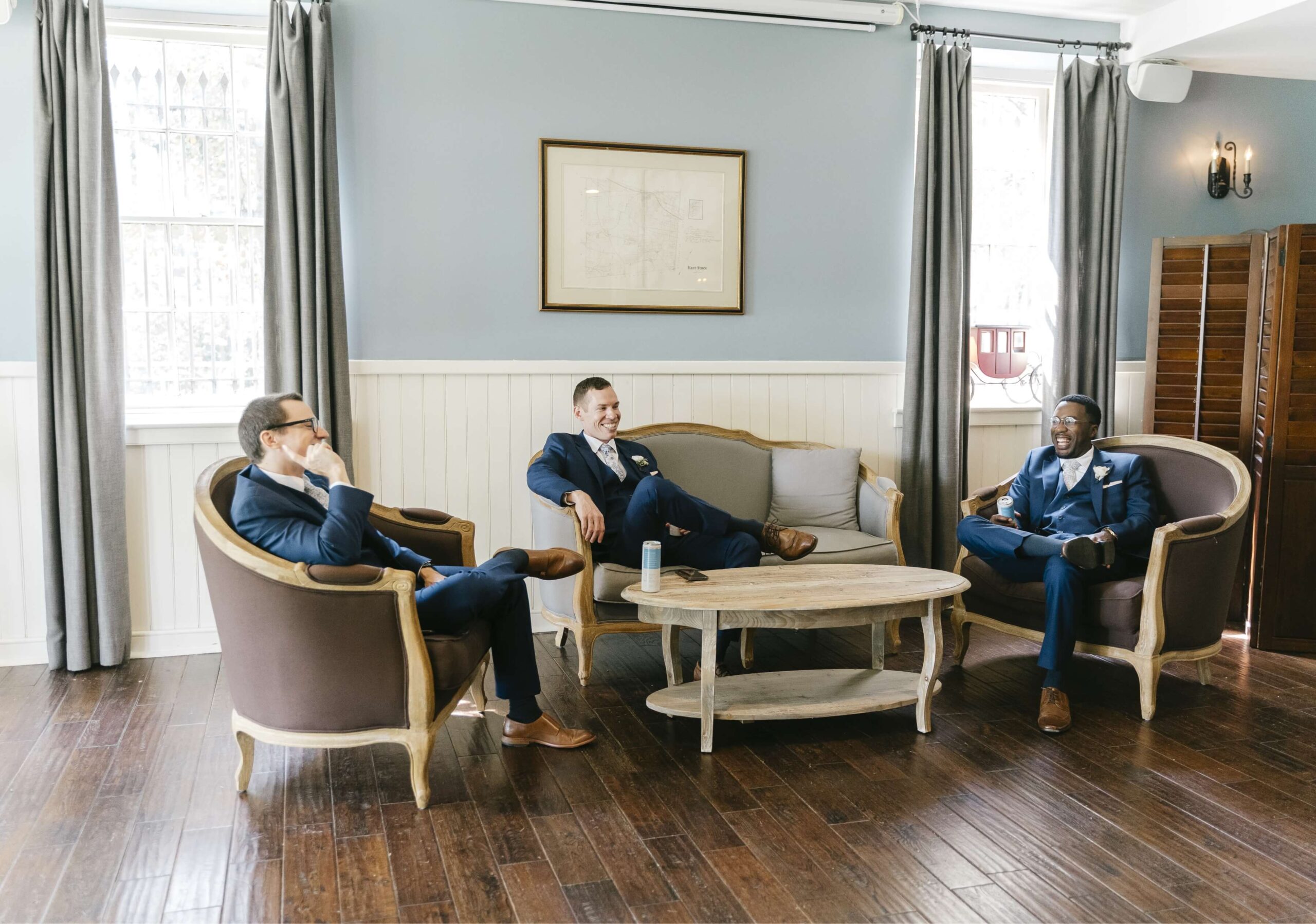 Three men in suits are sitting and talking on sofas around a coffee table in a room with wood flooring and blue-gray walls.