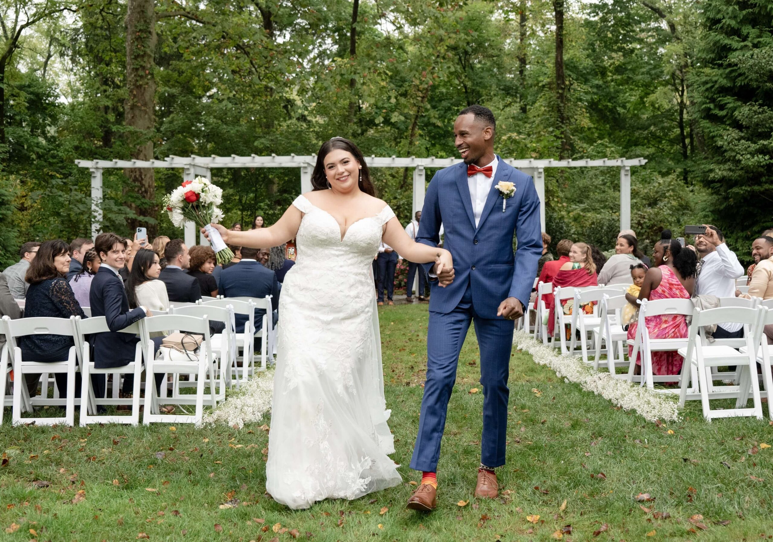 Bride in a white dress and groom in a blue suit walking down an outdoor aisle, smiling, with guests seated on either side.