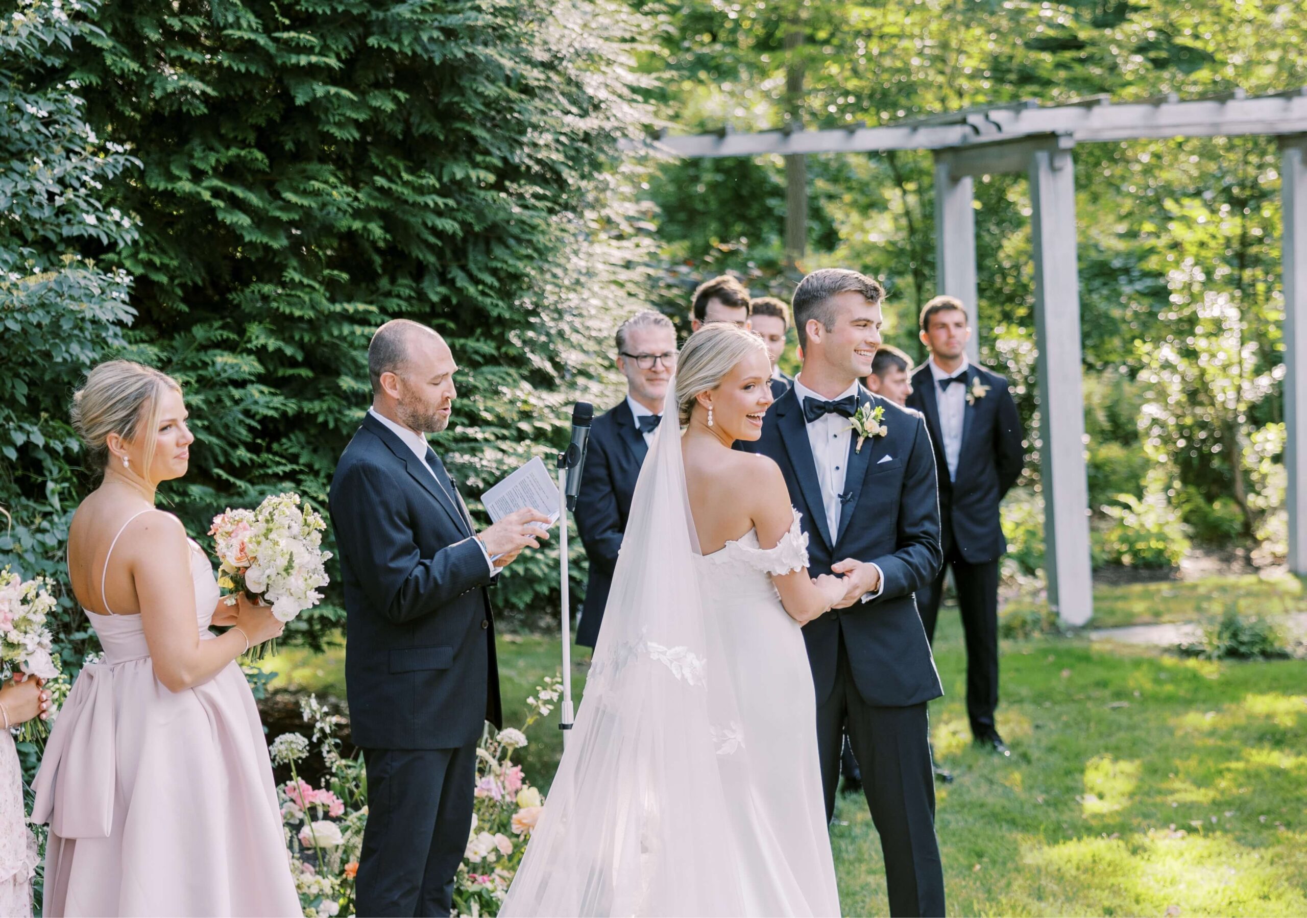 A bride and groom stand together holding hands outdoors under a trellis, surrounded by their wedding party.
