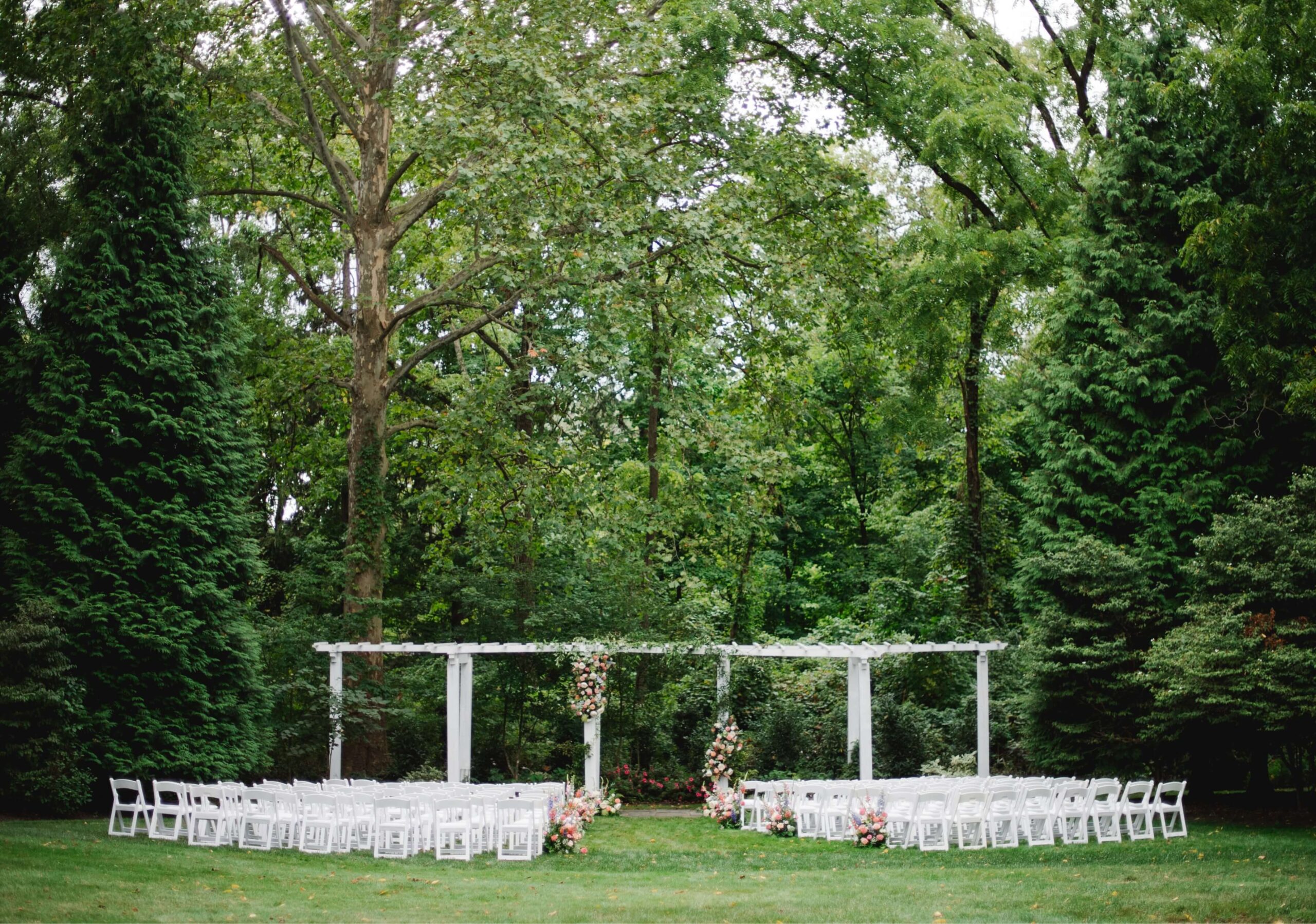 Outdoor wedding setup with white chairs arranged in rows facing a floral-decorated arbor, surrounded by tall green trees.