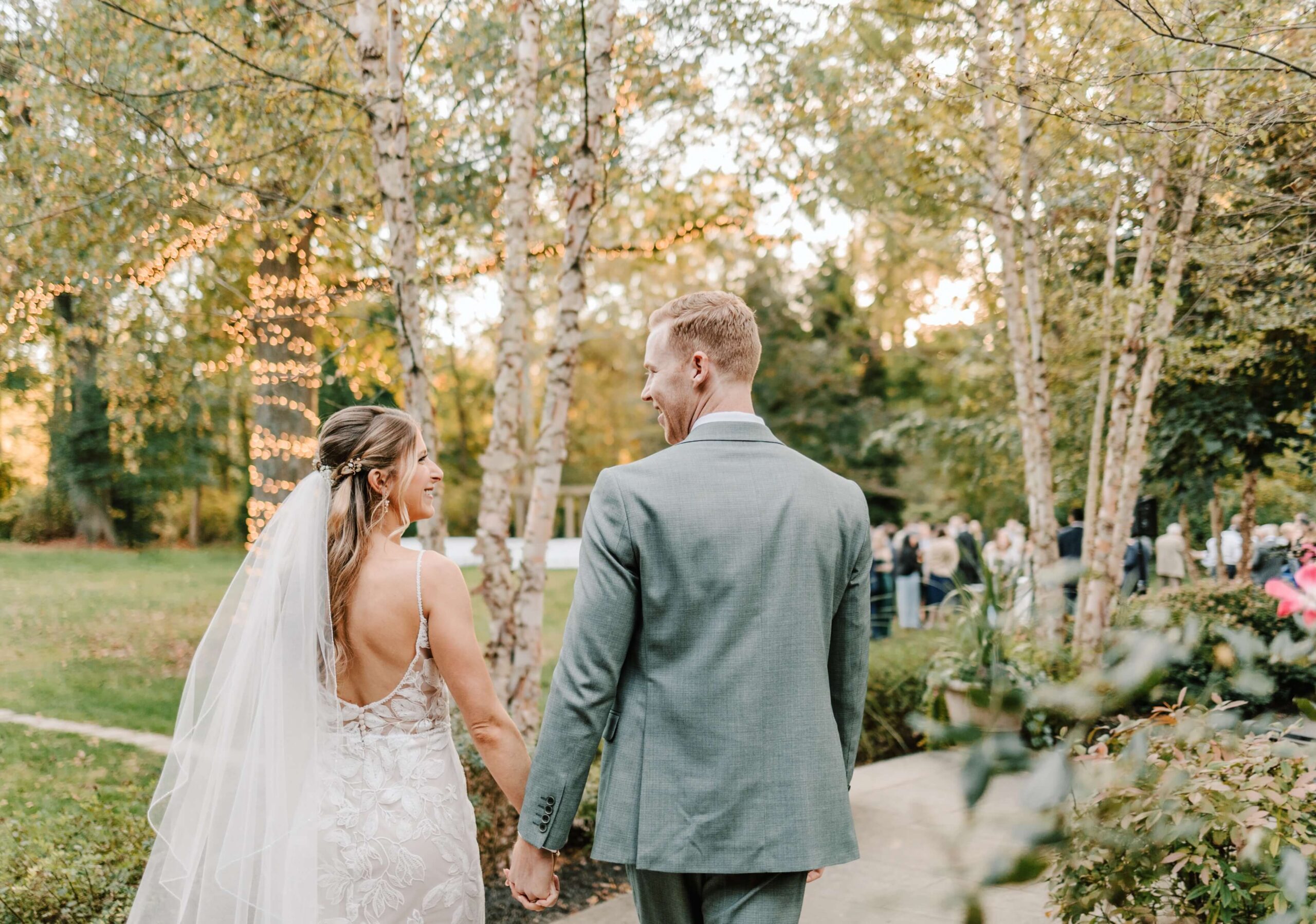 A bride and groom hold hands walking outdoors on a path lined with trees adorned with string lights. They are visible from the back in a lush garden setting.