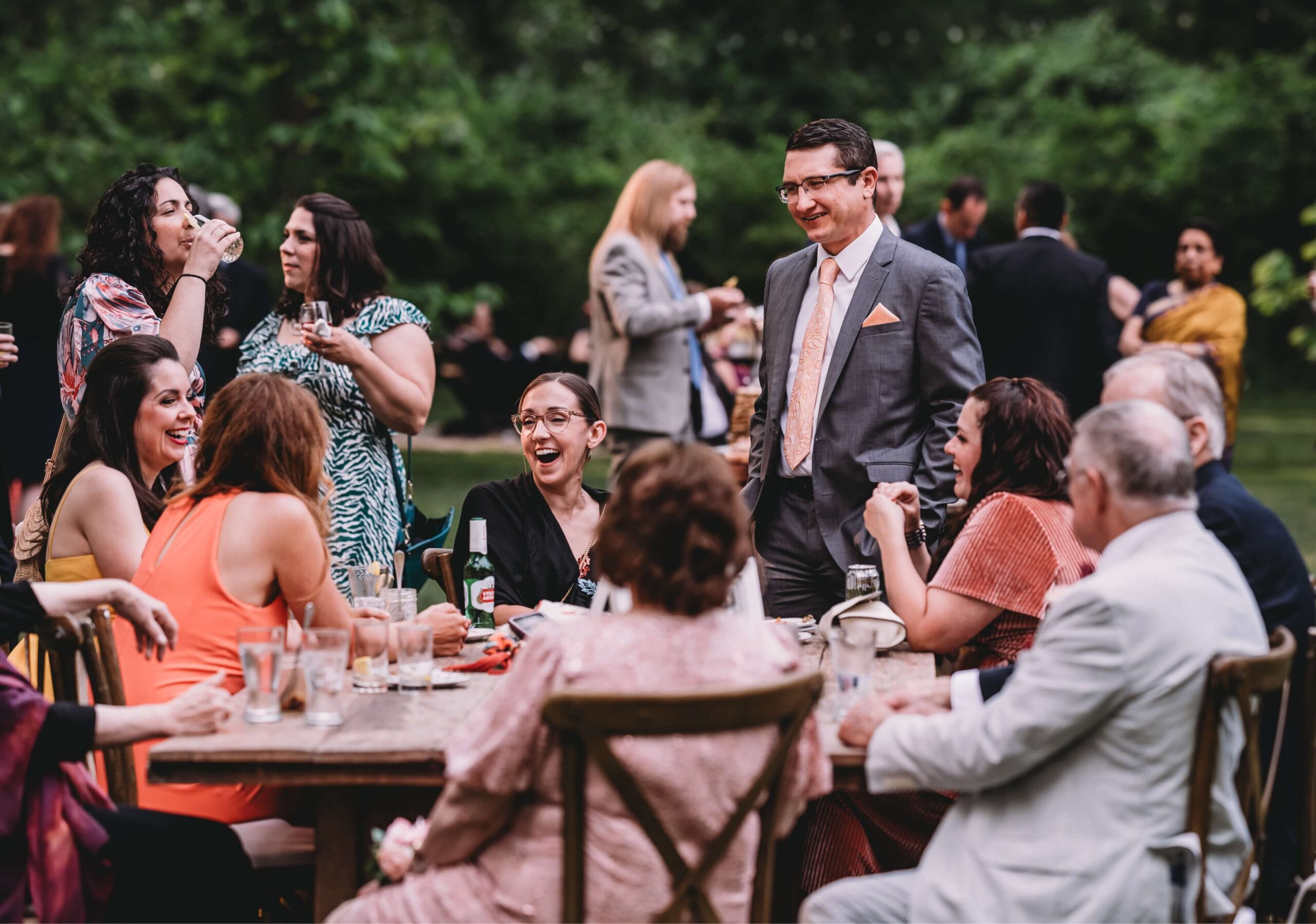 A group of people are gathered around a table outdoors, some sitting and others standing. They are engaged in conversation, with drinks in front of them. Trees are visible in the background.