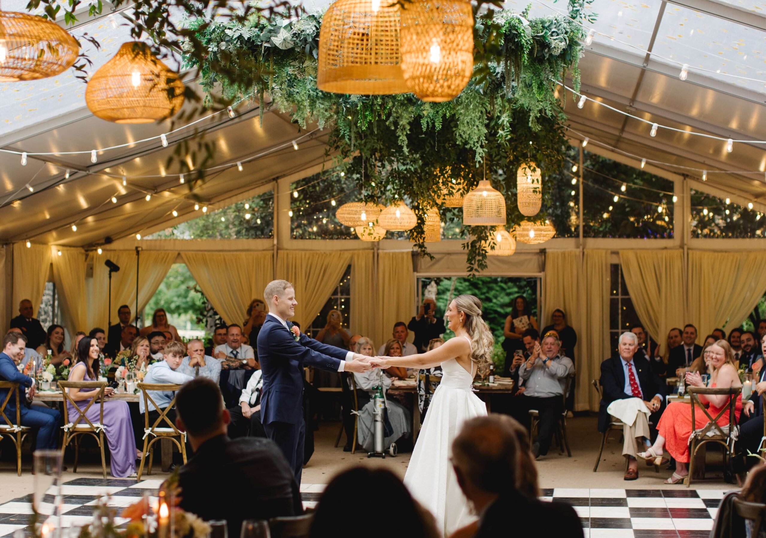 A couple dances on a checkered floor beneath a decorated tent with string lights, surrounded by seated guests.