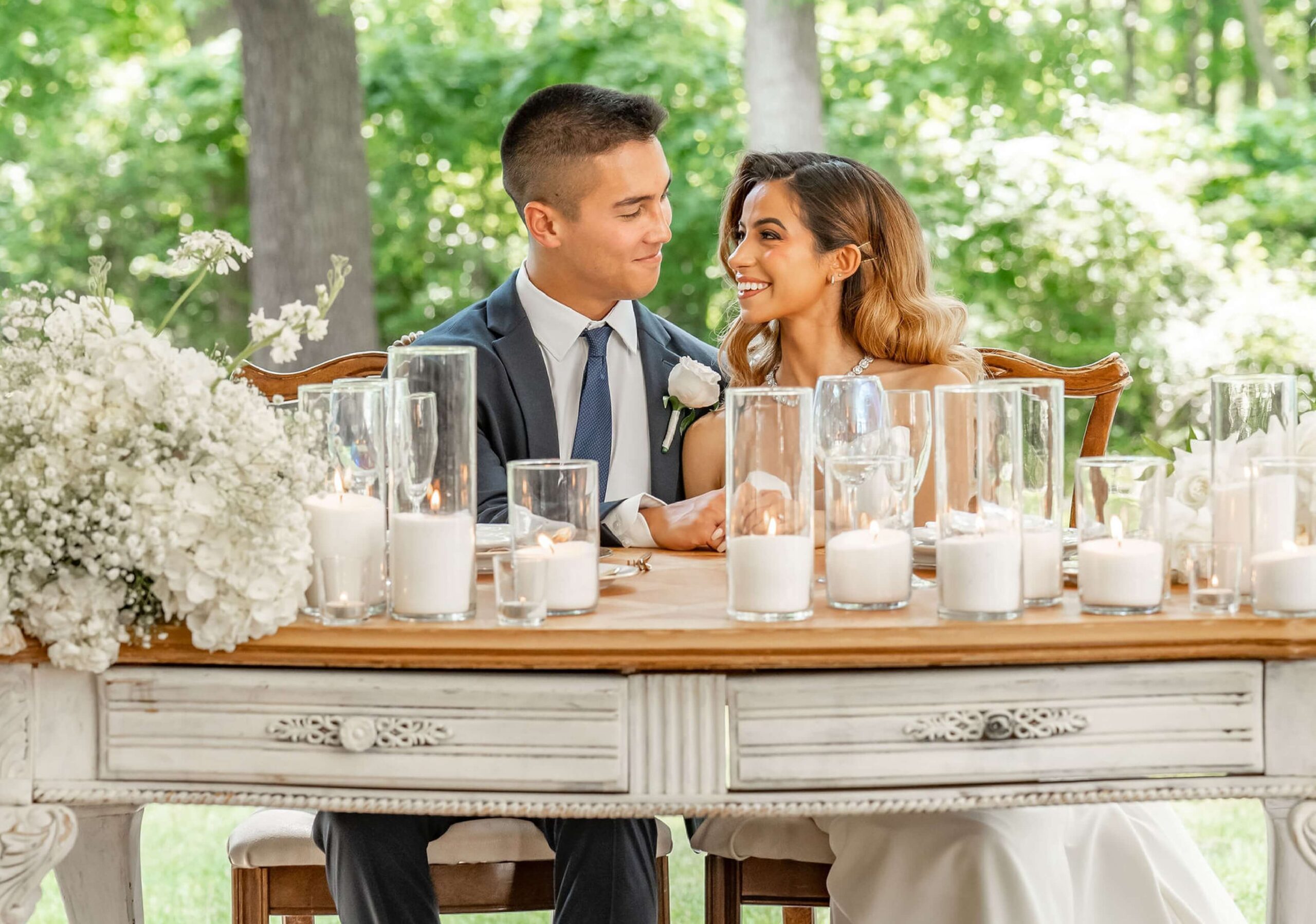 A couple in formal attire sits at a decorated wooden table outdoors, surrounded by white flowers and candles.