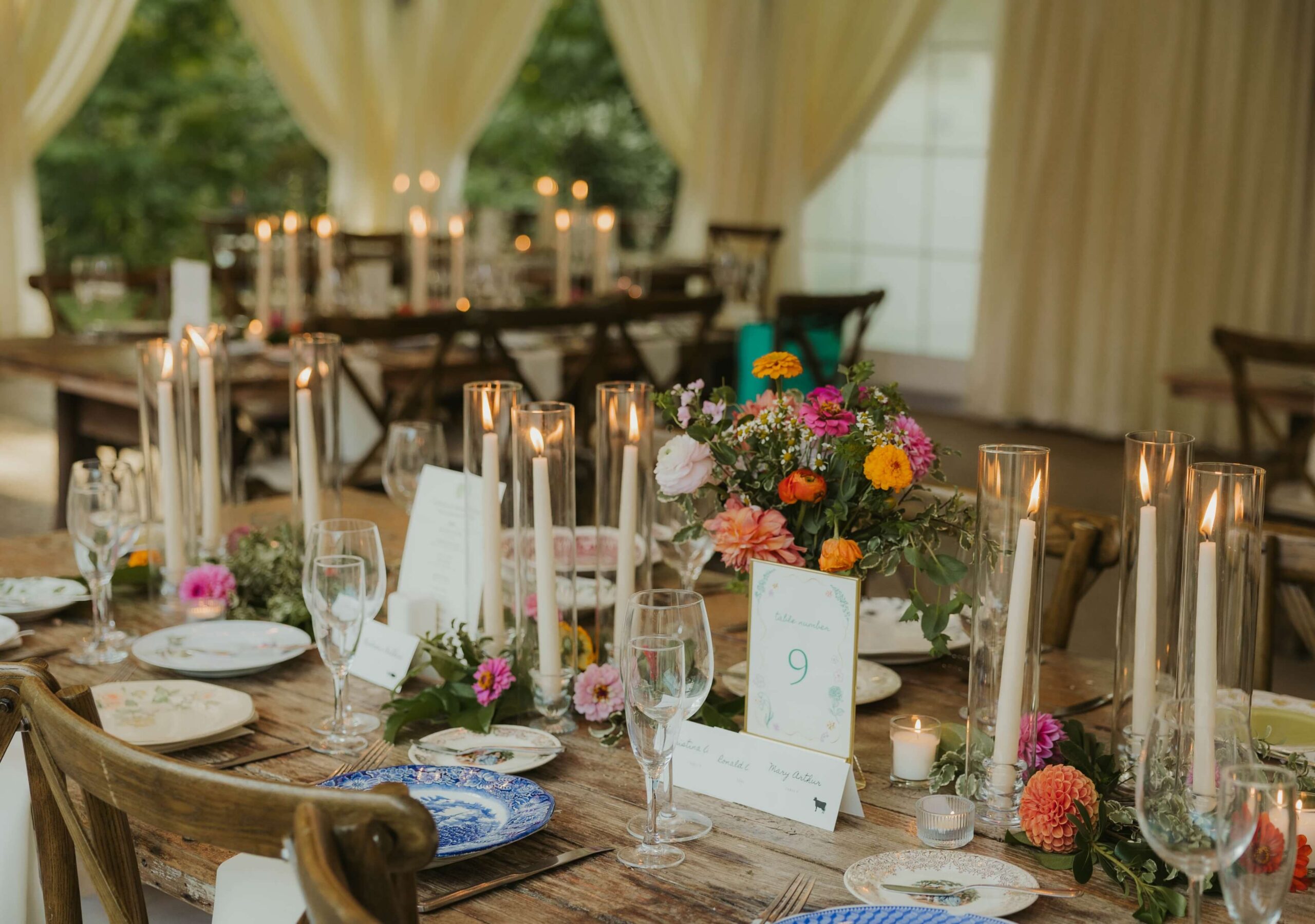 Elegant table setting with floral centerpieces, tall candles, numbered place cards, and assorted china plates on a wooden table under a draped canopy.