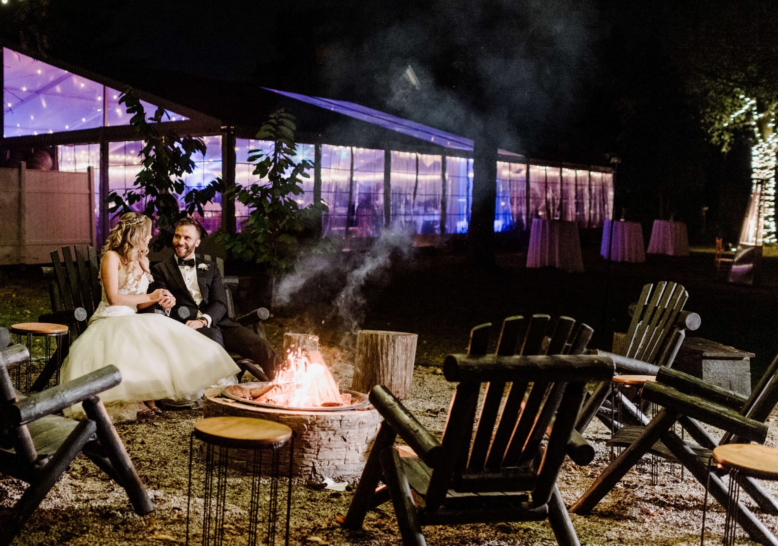 Bride and groom sitting by a lit fire pit at night, surrounded by empty wooden chairs, with an illuminated tent in the background.