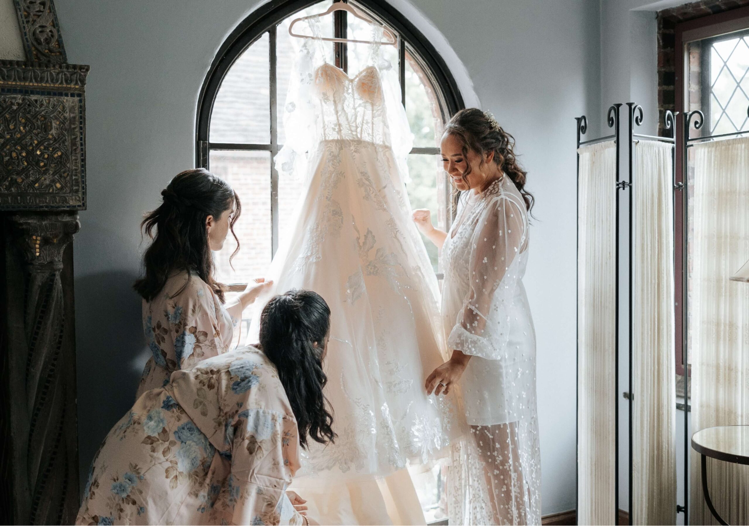 Three women admire a wedding dress hanging by a window in a softly lit room. They are wearing light, floral robes.