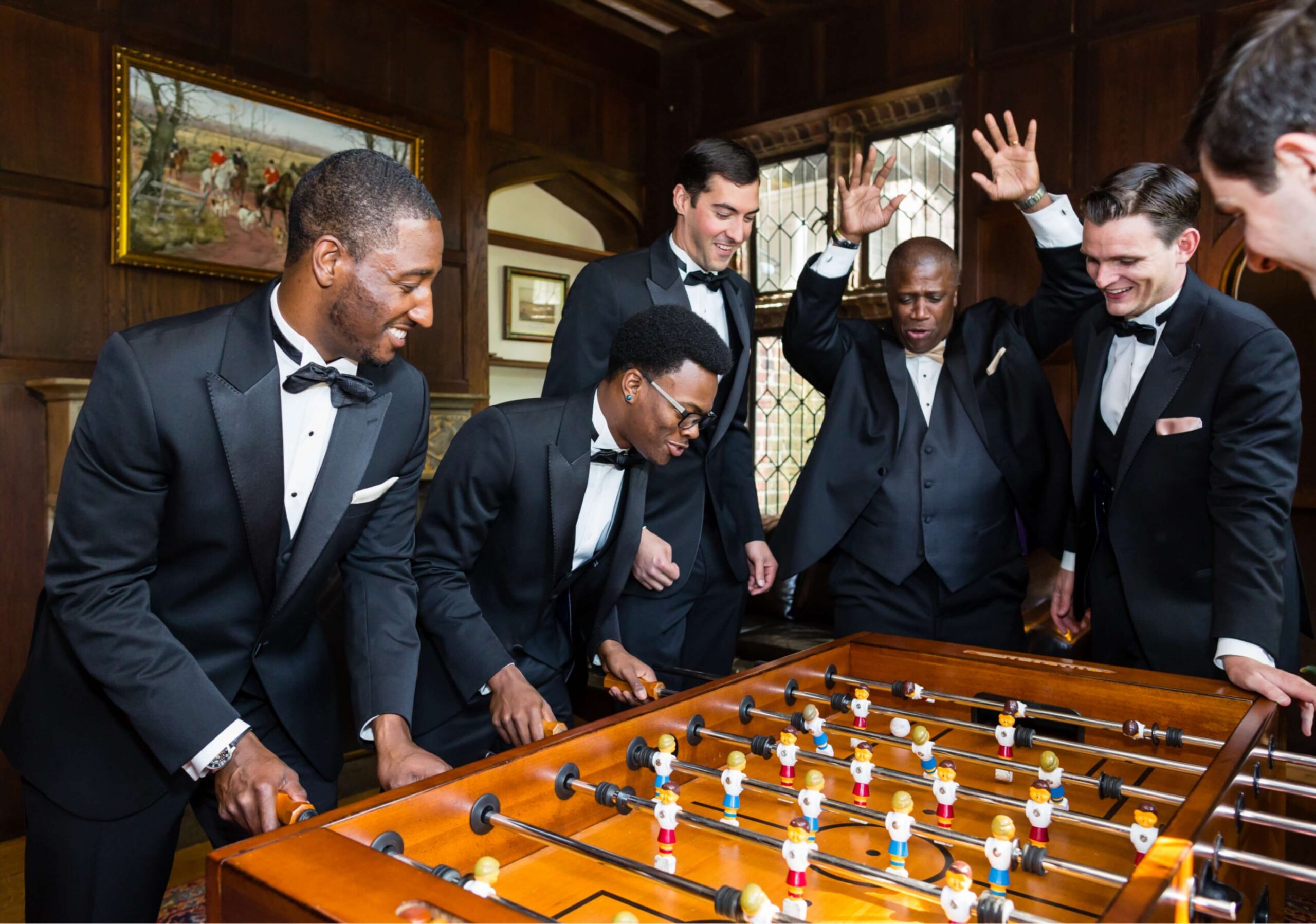 Five men in formal wear play a lively game of foosball in a wood-paneled room, with some raising their hands in excitement.