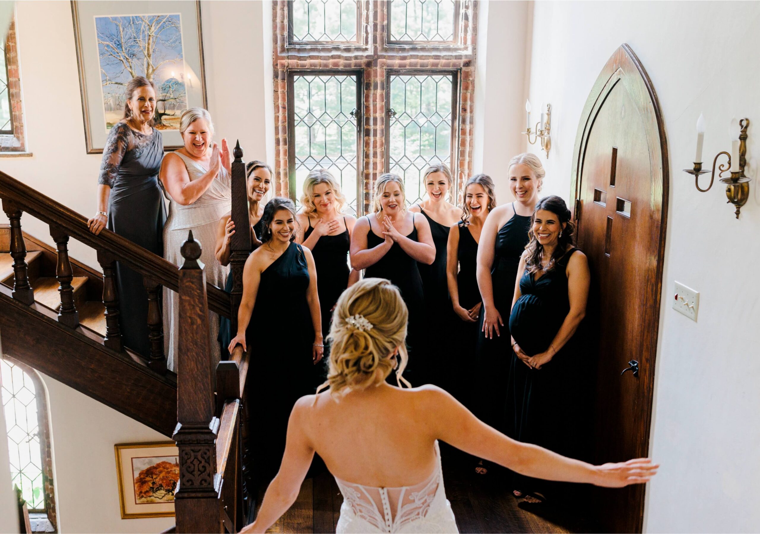 A bride in a white dress stands at the bottom of a staircase, facing a group of bridesmaids who are reacting with excitement and surprise.