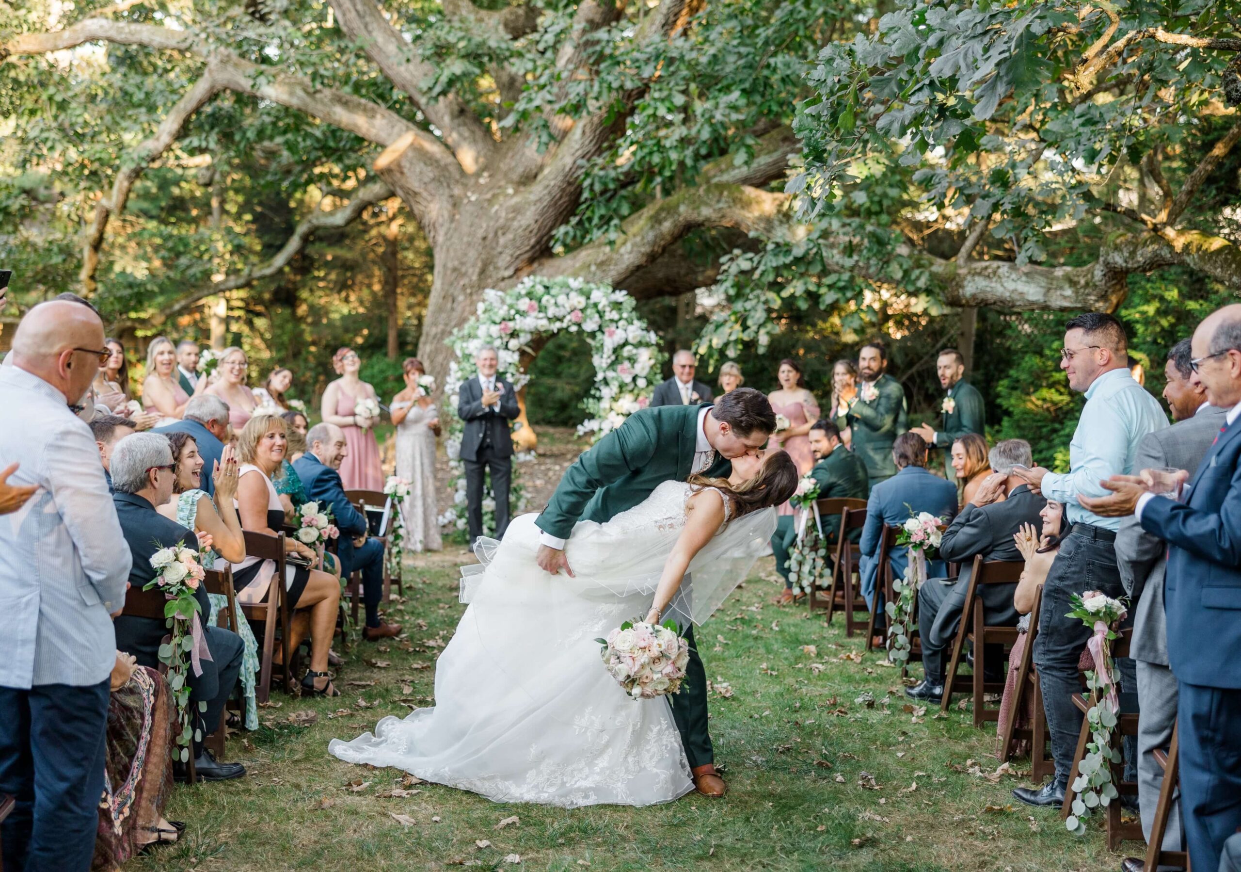Couple kissing during their wedding ceremony outdoors, surrounded by guests and greenery, with a floral arch in the background.
