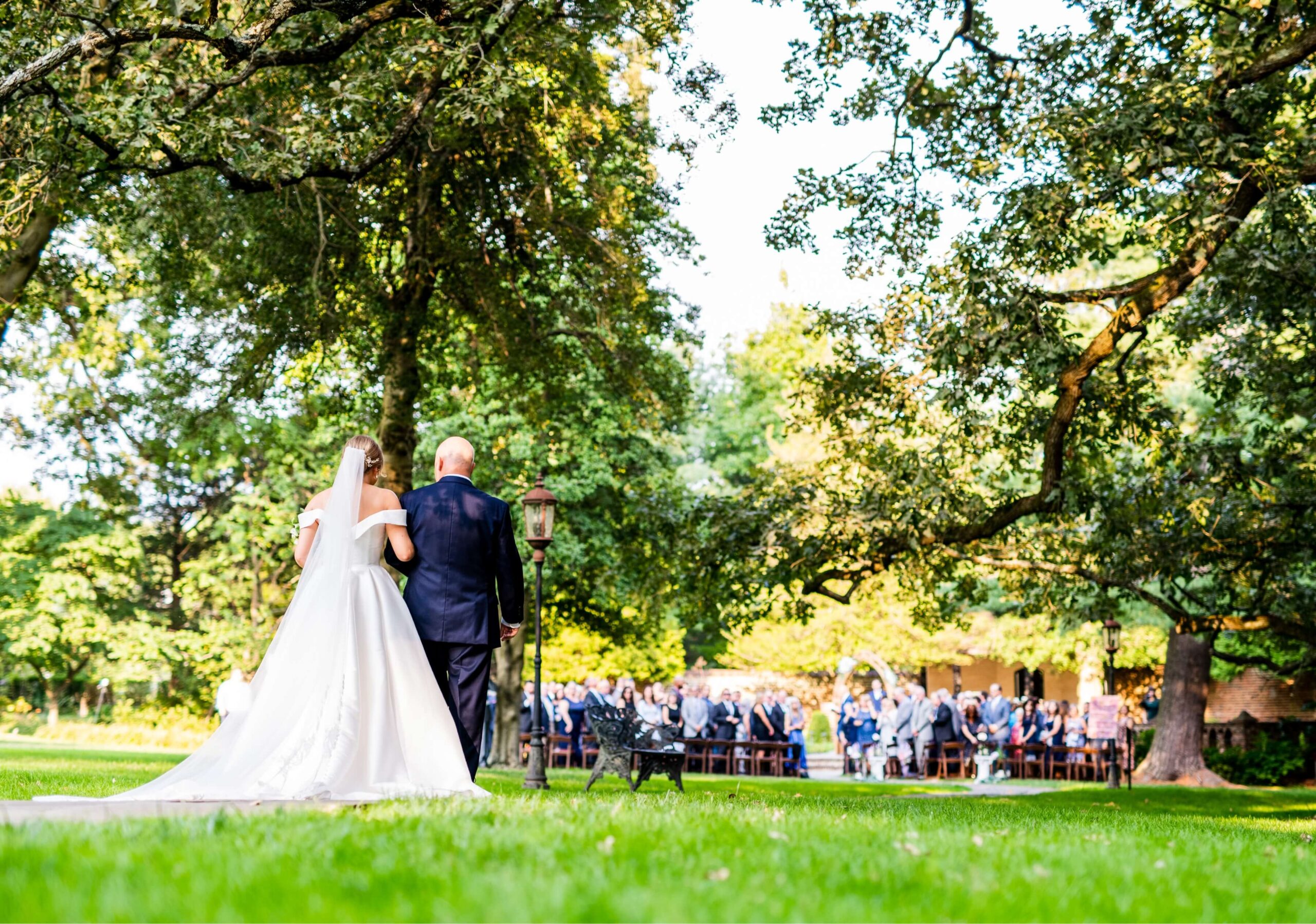 A bride and an older man walk down a grassy aisle outdoors, approaching a seated crowd under large trees in a sunny setting.