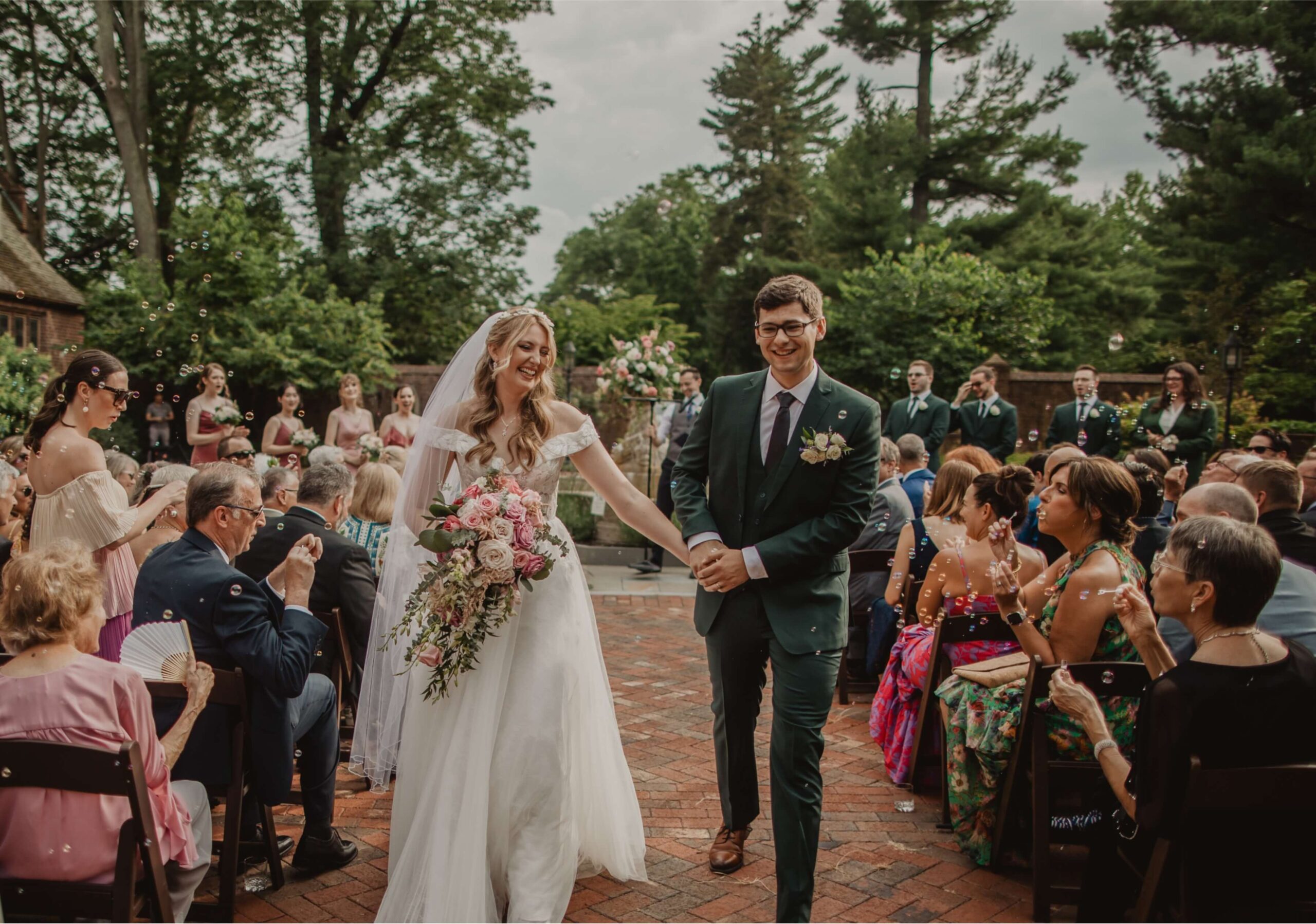 A bride and groom walk down the aisle outdoors, smiling and holding hands. Guests seated on either side applaud. The bride holds a bouquet, and trees are visible in the background.