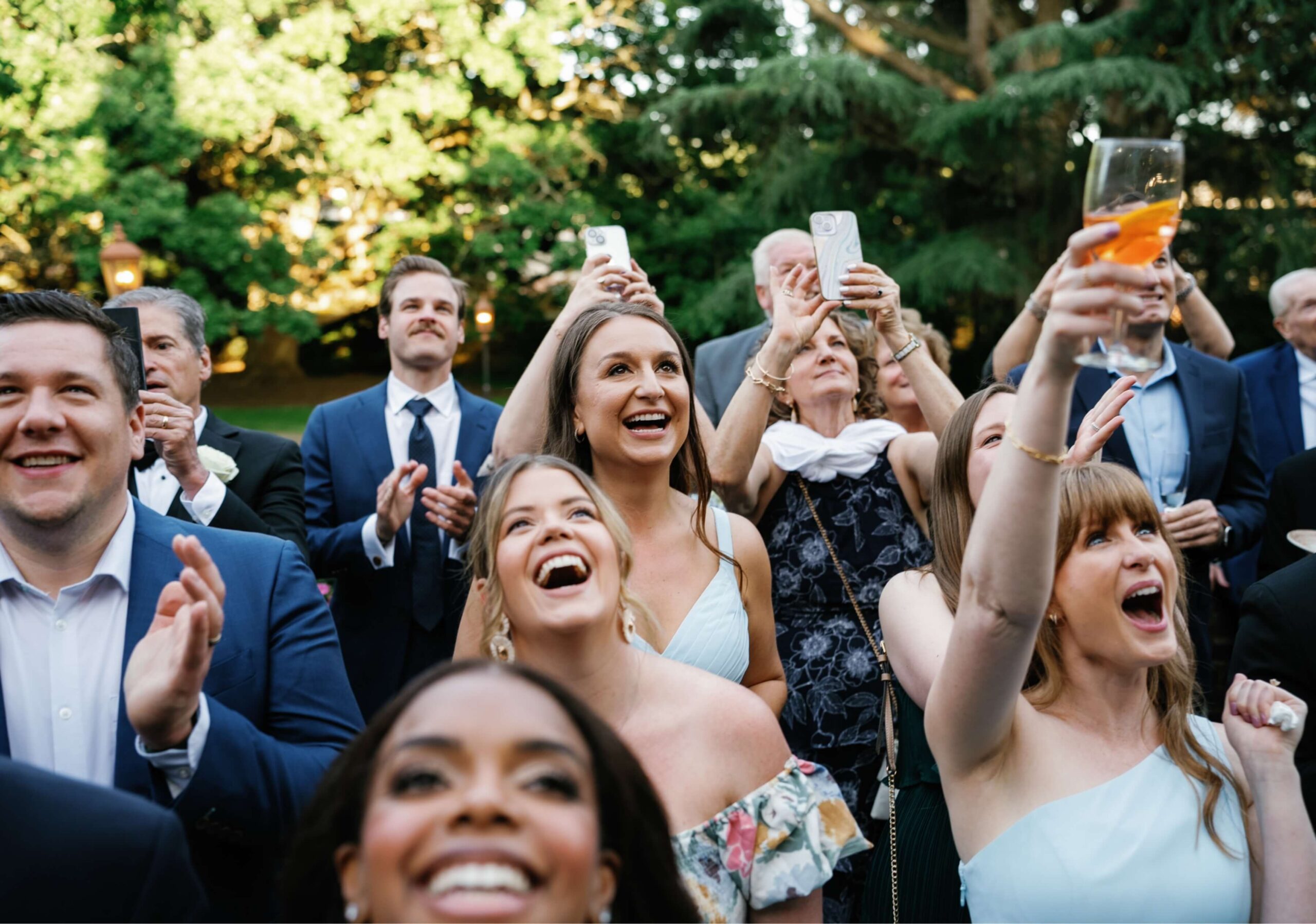 A group of people outdoors, dressed formally, smiling and cheering while holding drinks and phones.