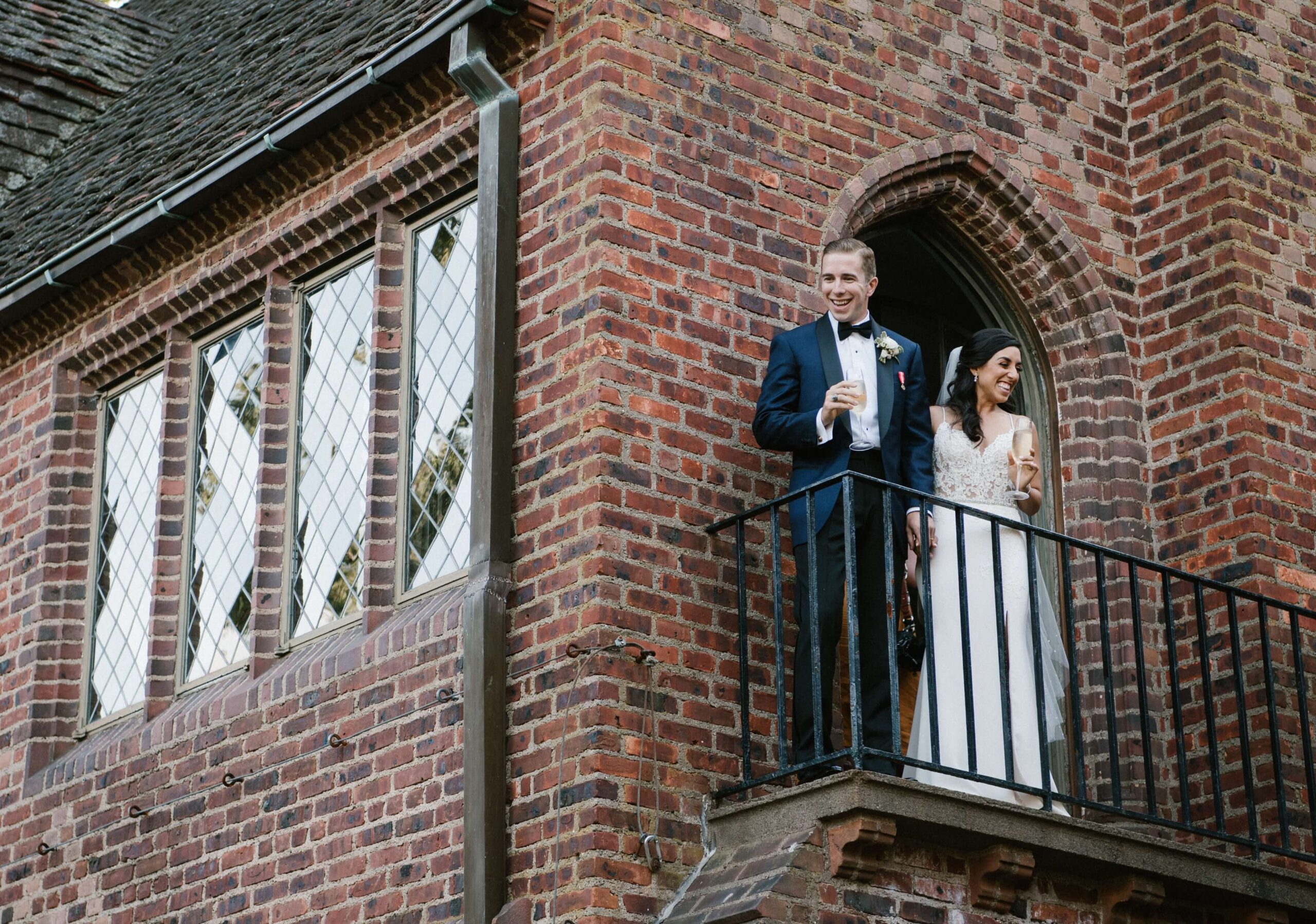 A couple in formal attire stands on a brick building balcony, looking out and smiling.
