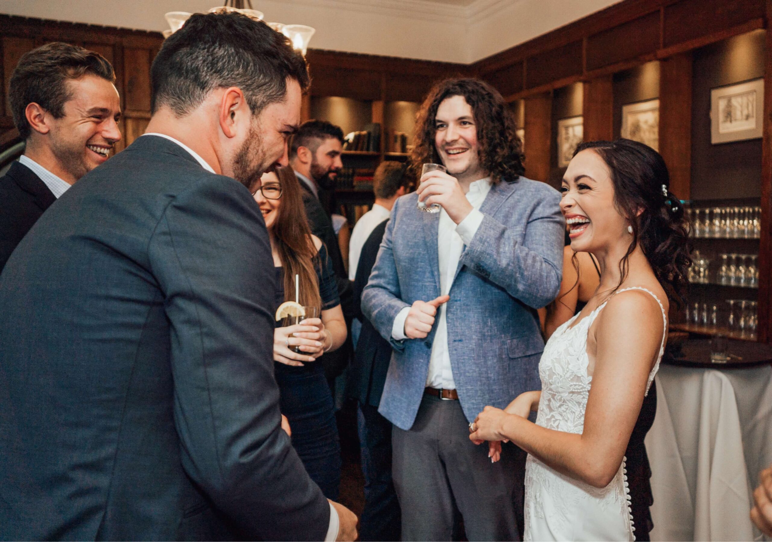 A bride in a white dress and a group of people in formal attire are smiling and talking in a room with wooden paneling and framed pictures.