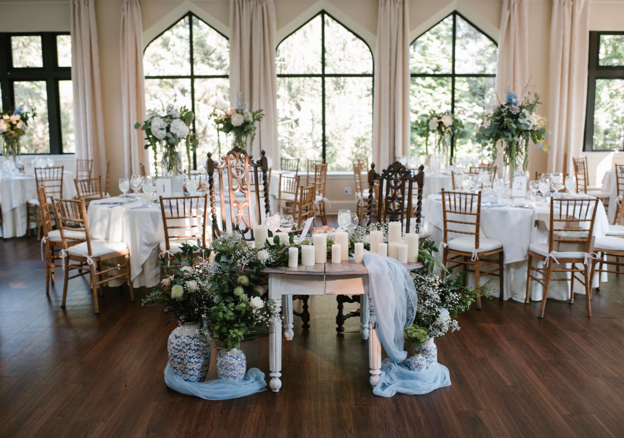 Elegant wedding reception setup with a table adorned with candles and flowers. Wooden chairs surround round tables with white linens, set in a room with large arched windows.