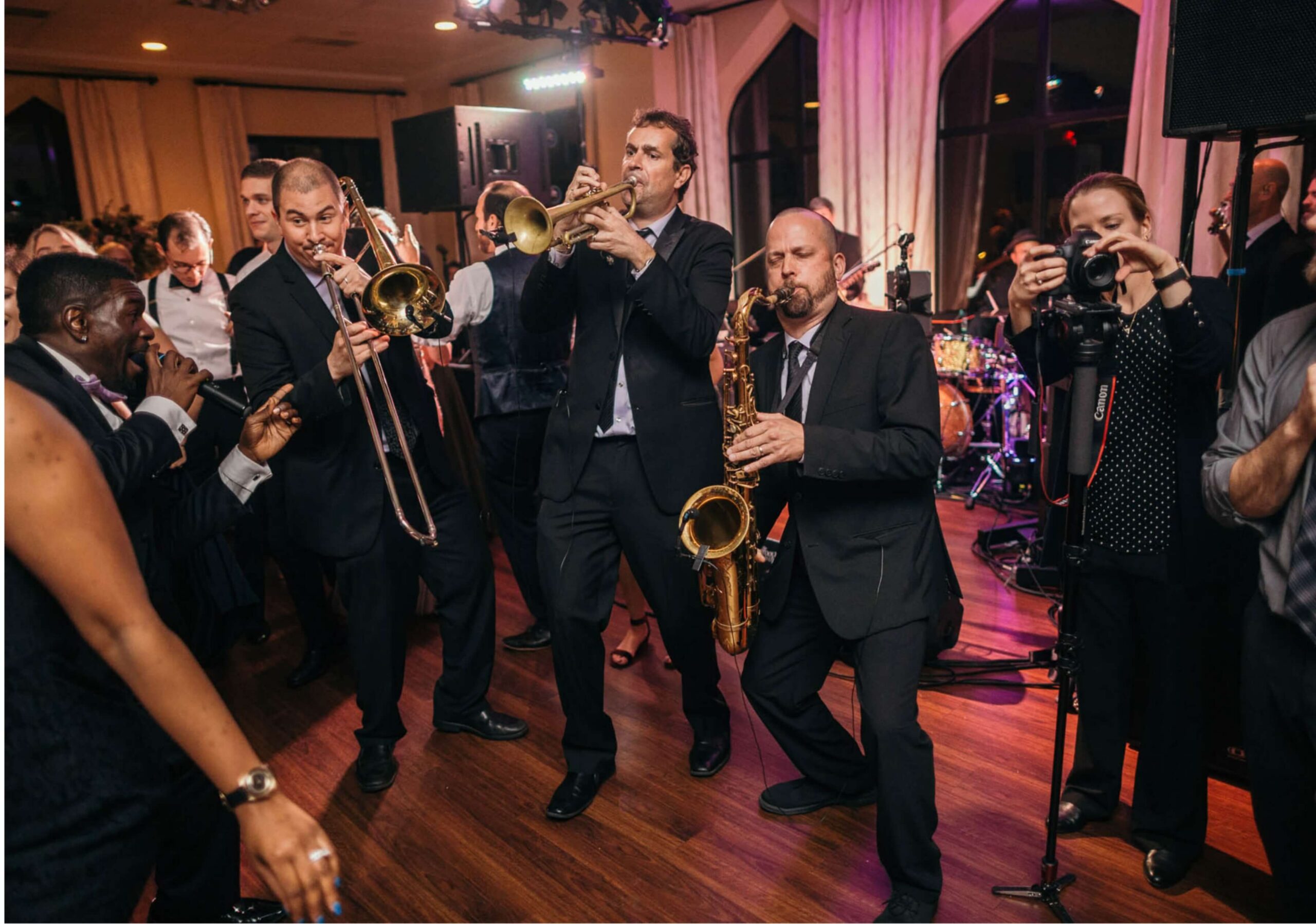 Musicians in formal wear play trombones, trumpet, and saxophone at an indoor event, surrounded by an audience, including a photographer capturing the moment.