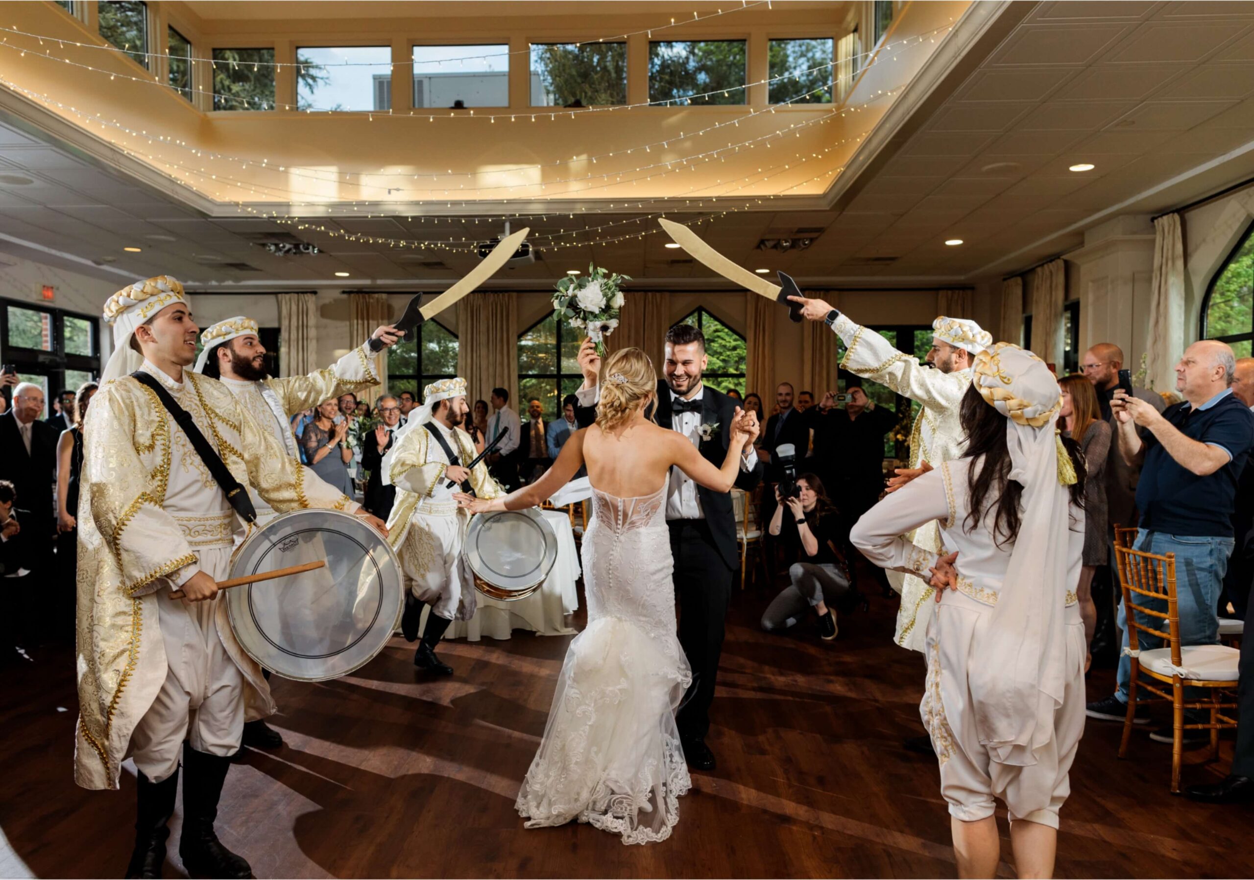 Bride and groom dance surrounded by performers in traditional attire holding swords, with guests watching in an elegant reception hall.