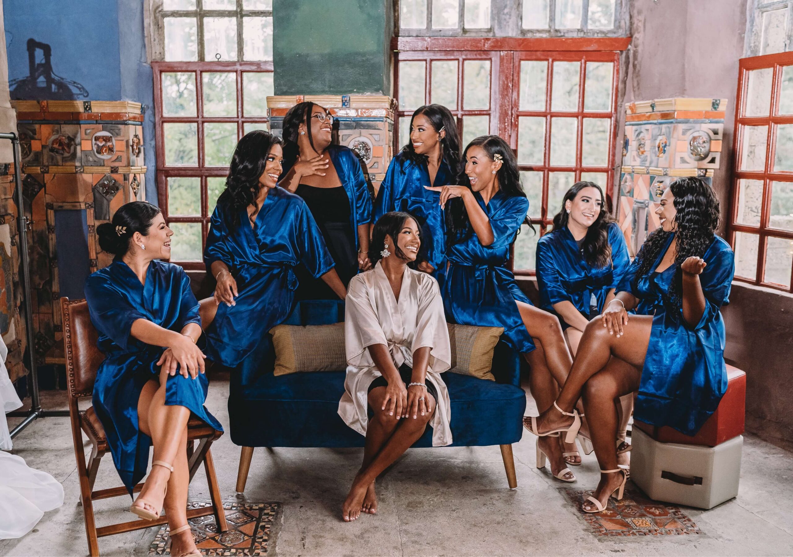 A group of eight women in blue and white robes sit and stand around a blue couch in a room with large windows, smiling and interacting with each other.