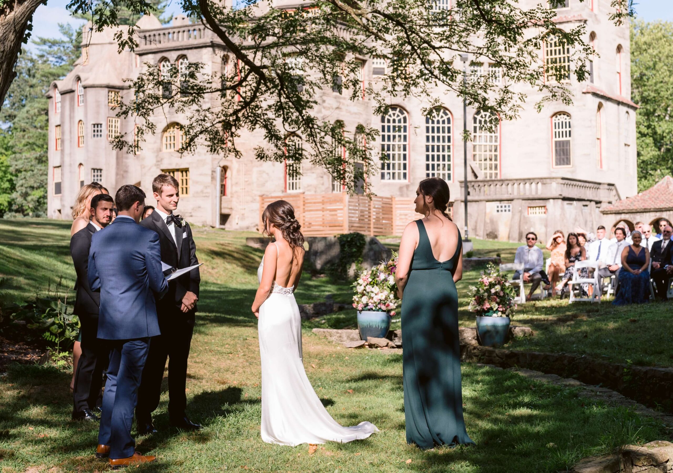 Outdoor wedding ceremony at a historic estate. A couple stands facing each other with an officiant, bridesmaid, and guests seated in the background.
