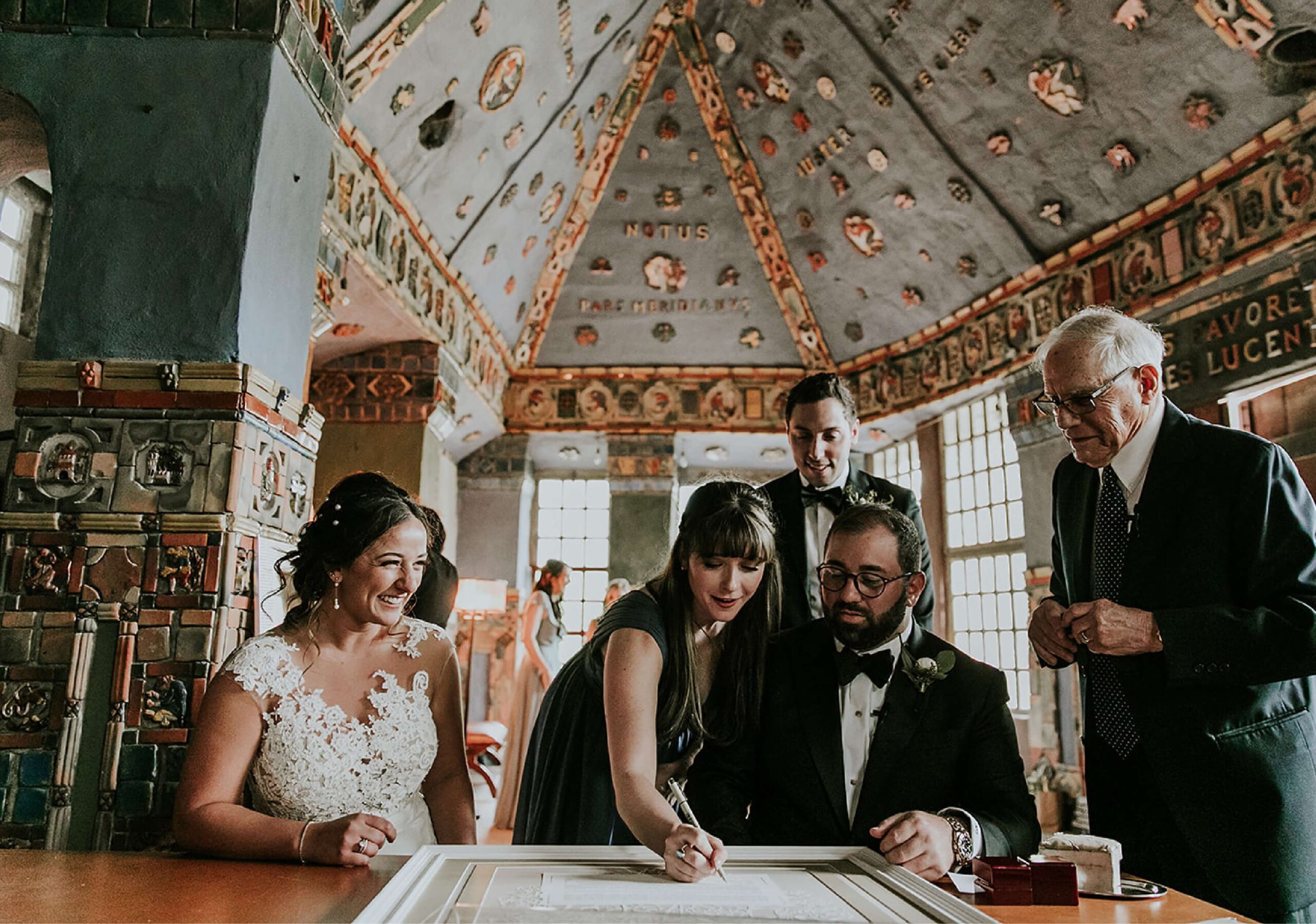 Bride and groom observe as a woman signs a document in an intricately decorated room, with two men standing nearby.