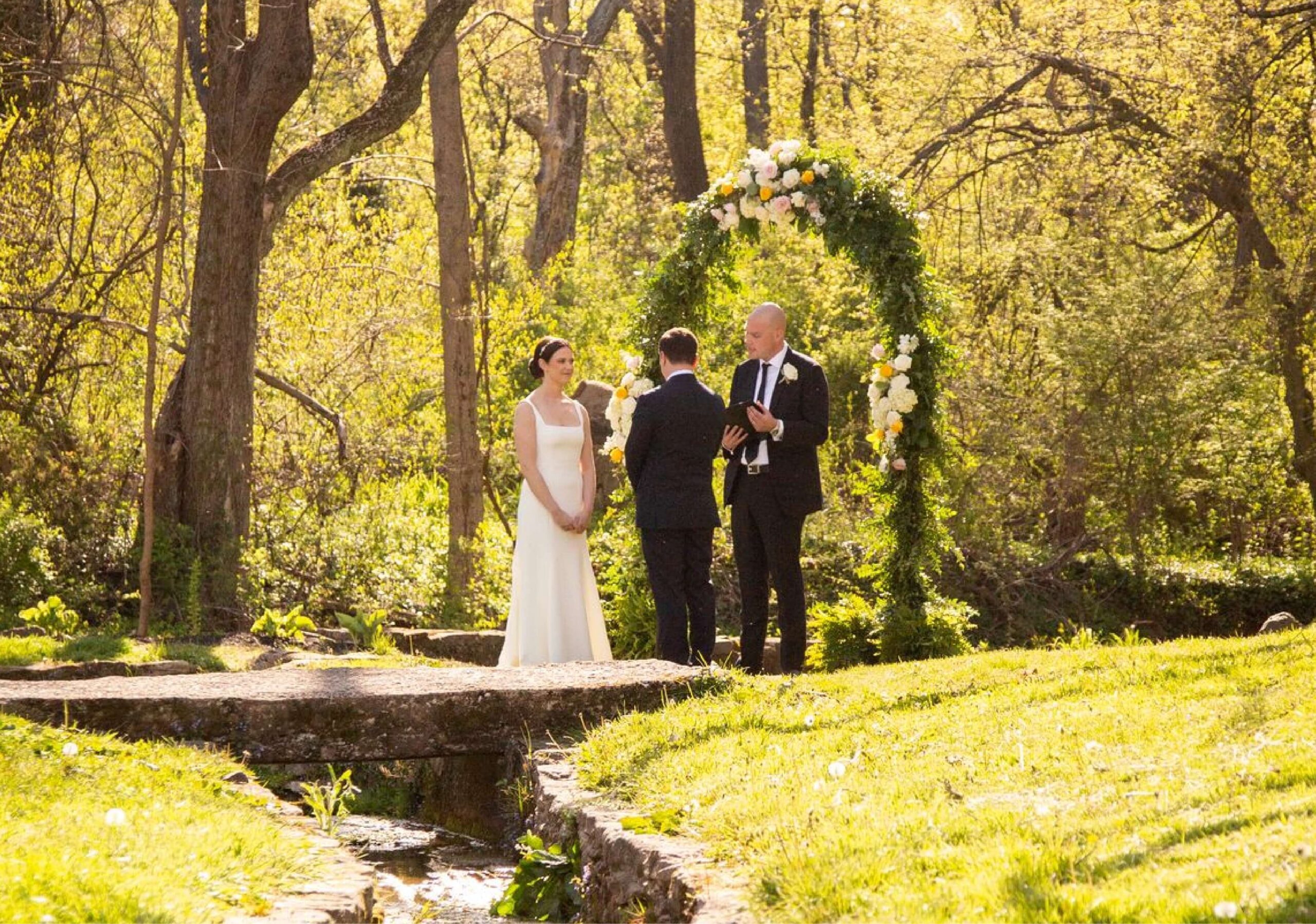 A bride and groom stand beside an officiant under a floral arch in a wooded outdoor setting, the sun filtering through the trees.
