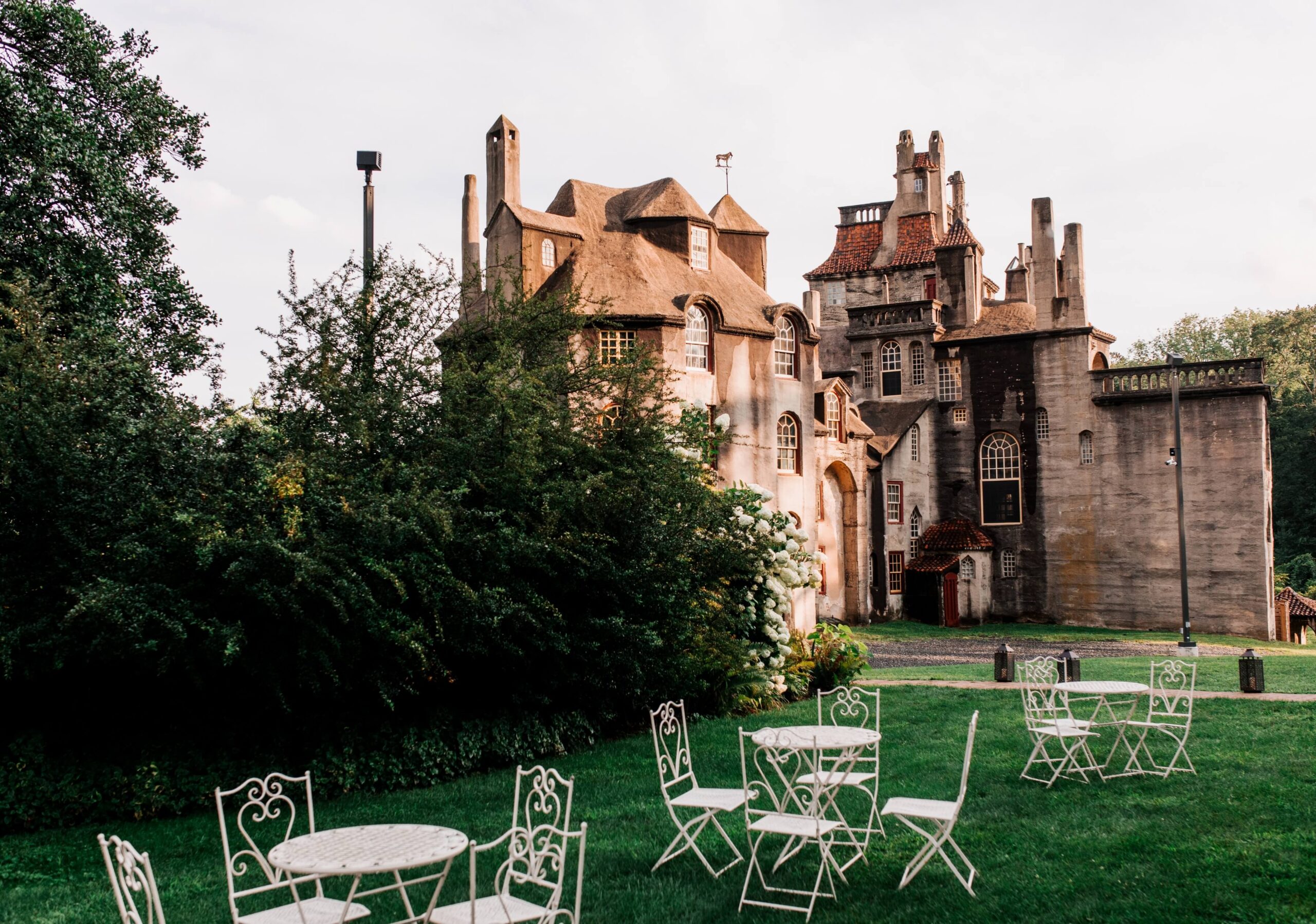 A whimsical stone building with unique architecture stands amidst greenery. White metal tables and chairs are placed on a grassy area in the foreground.