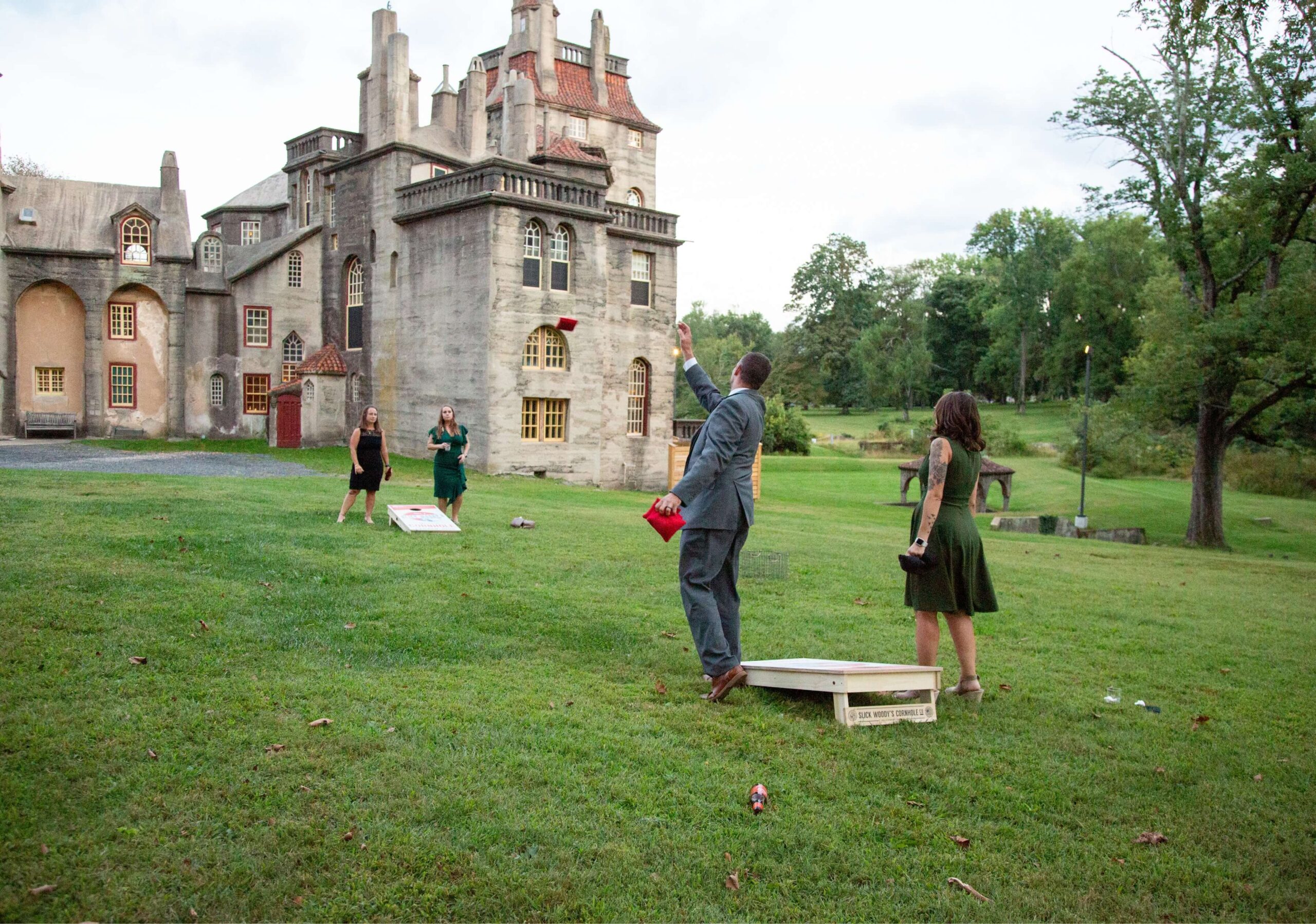 People playing cornhole on grass in front of a large stone building on a cloudy day.