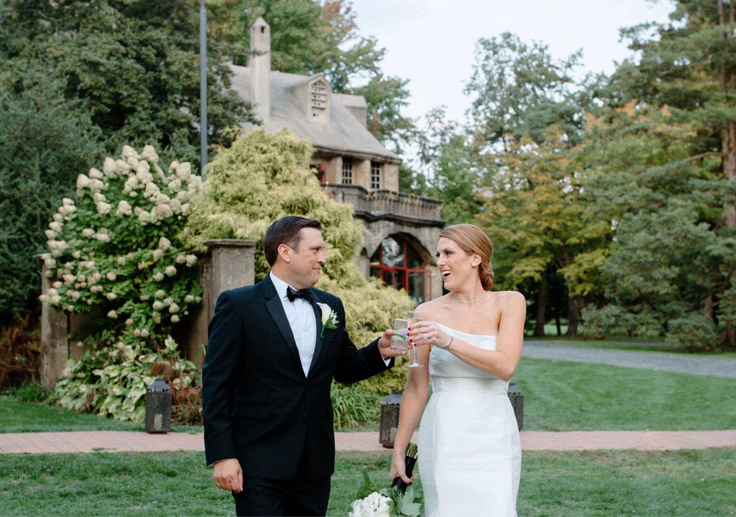 A man in a tuxedo and a woman in a white dress hold glasses, standing outdoors with trees and a cottage-like building in the background.
