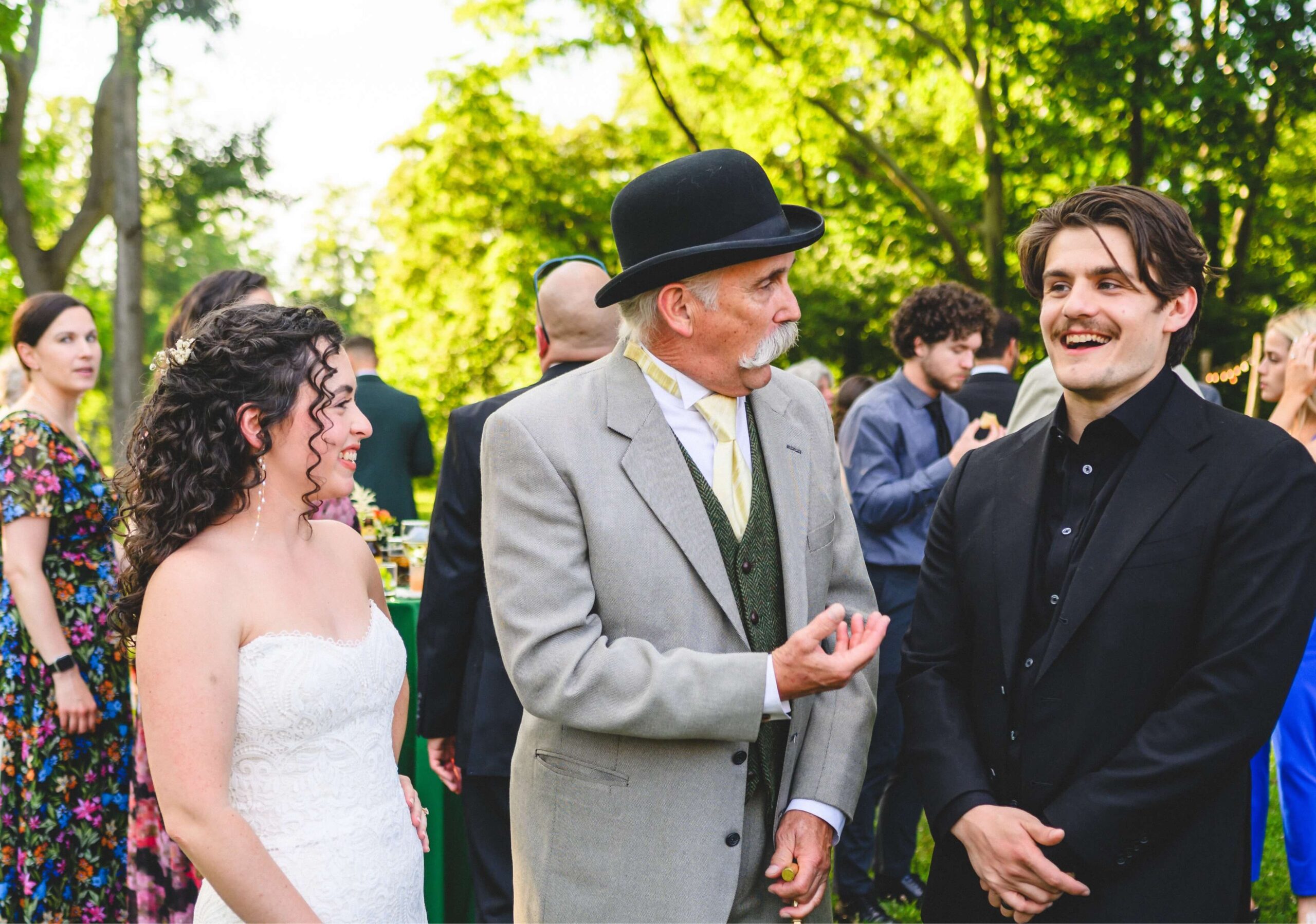 A bride, an older man in a bowler hat, and a man in a black suit chat at an outdoor gathering with other guests in the background.