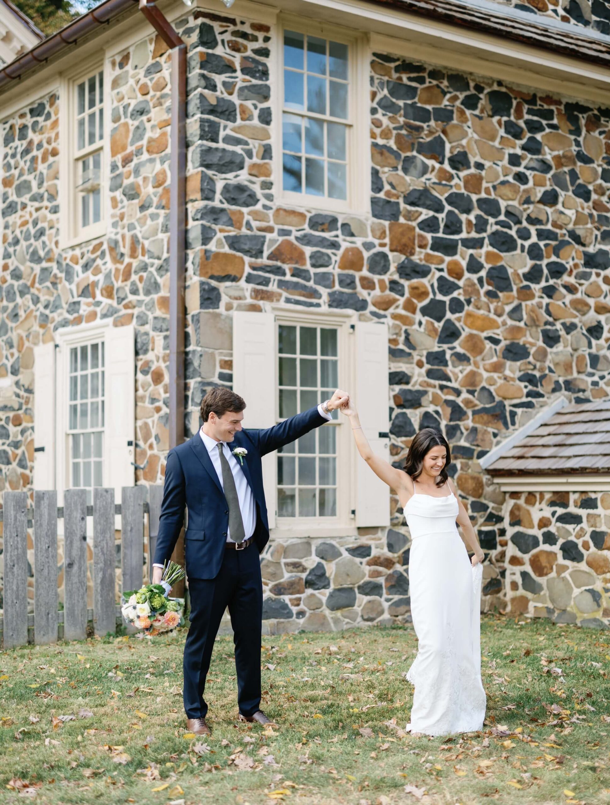 A couple in formal attire dances joyfully outside a stone house. The man holds a bouquet, and they are surrounded by grass and fallen leaves.