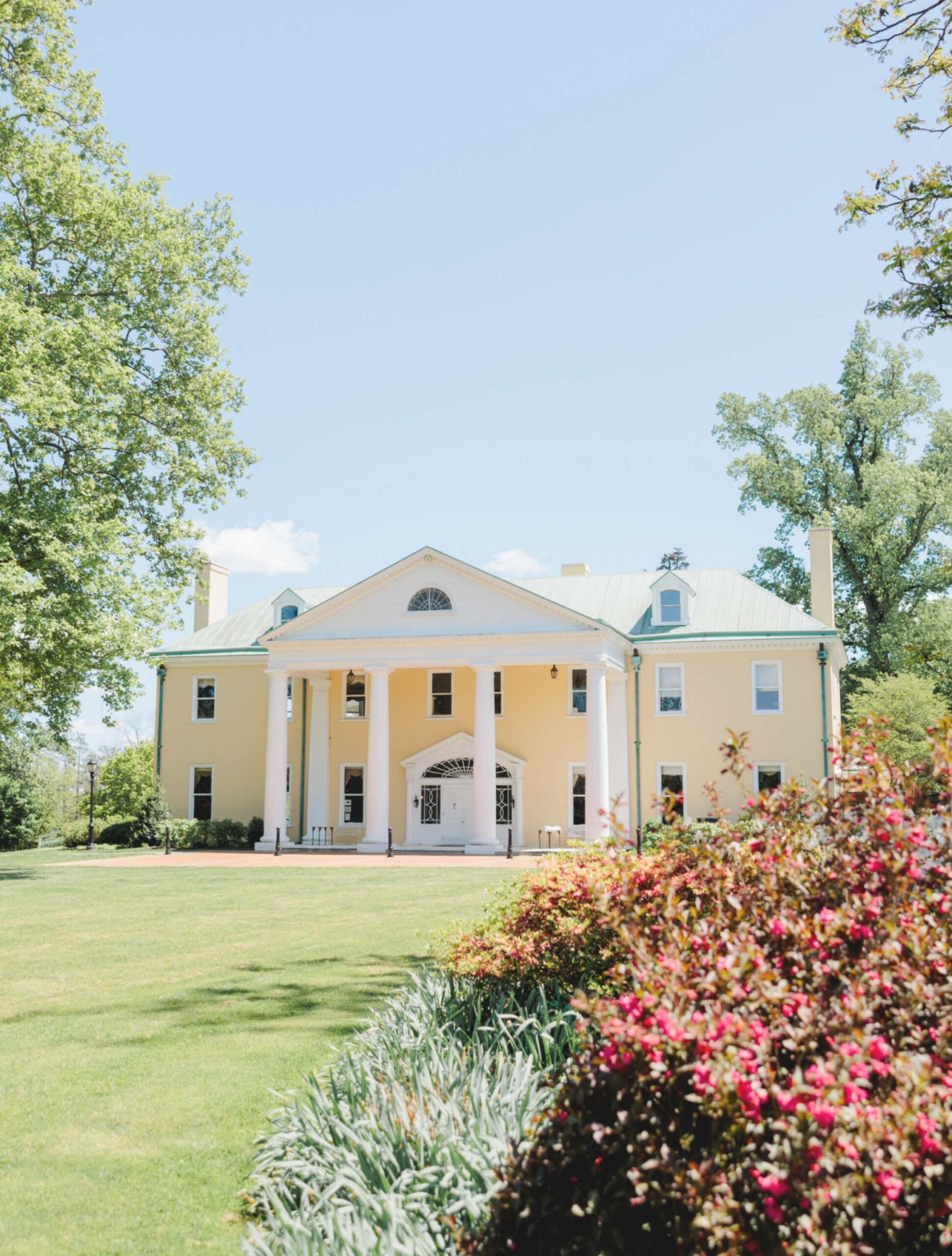 A large yellow mansion with white columns stands amidst a well-kept lawn and colorful flowerbeds, surrounded by tall trees under a clear blue sky.