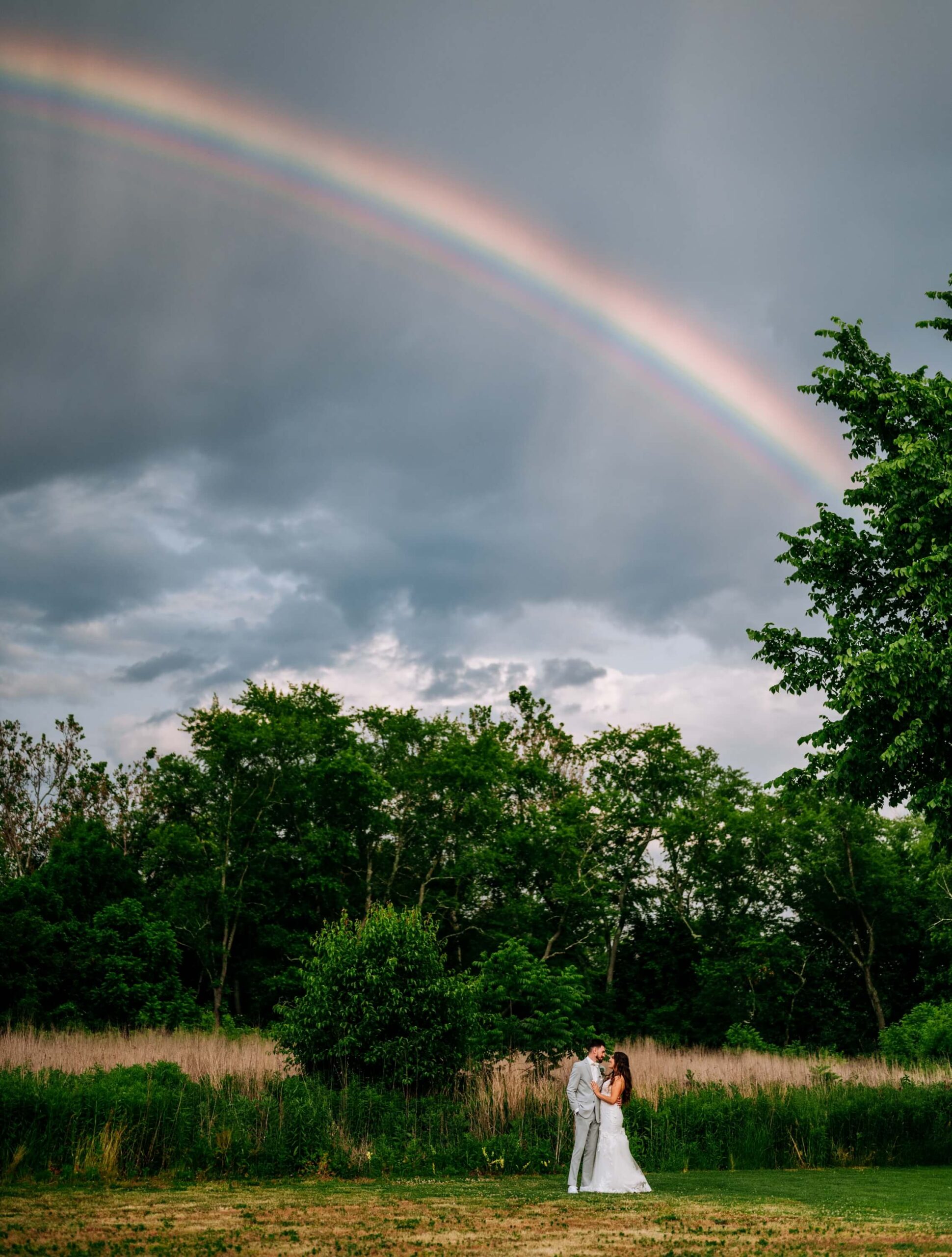 A couple stands embracing in a grassy field under a cloudy sky with a vivid rainbow arching overhead. Trees are in the background.