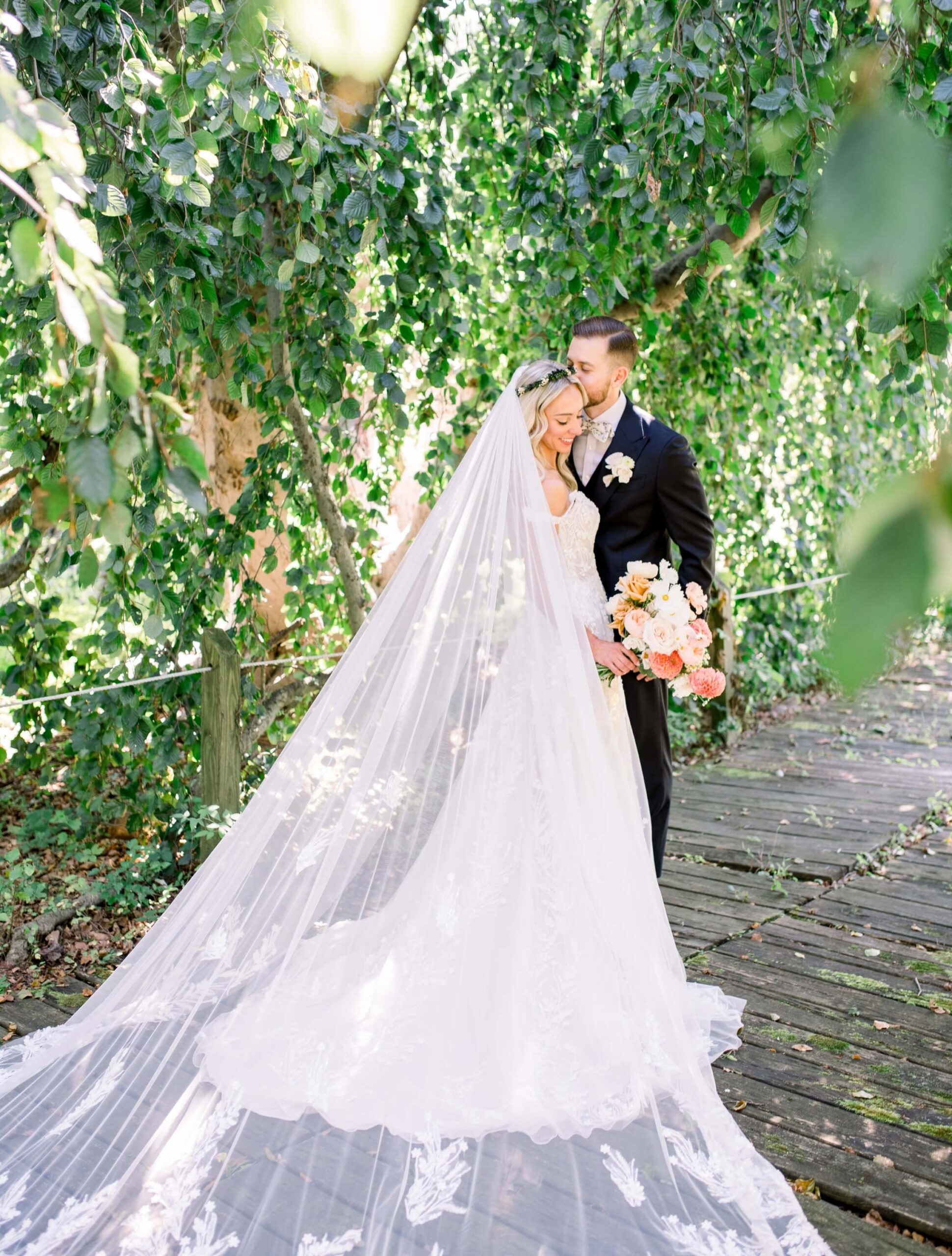 Bride and groom standing on a wooden path under lush greenery. The bride wears a long white gown with a lace veil, holding a bouquet. The groom is in a black suit.