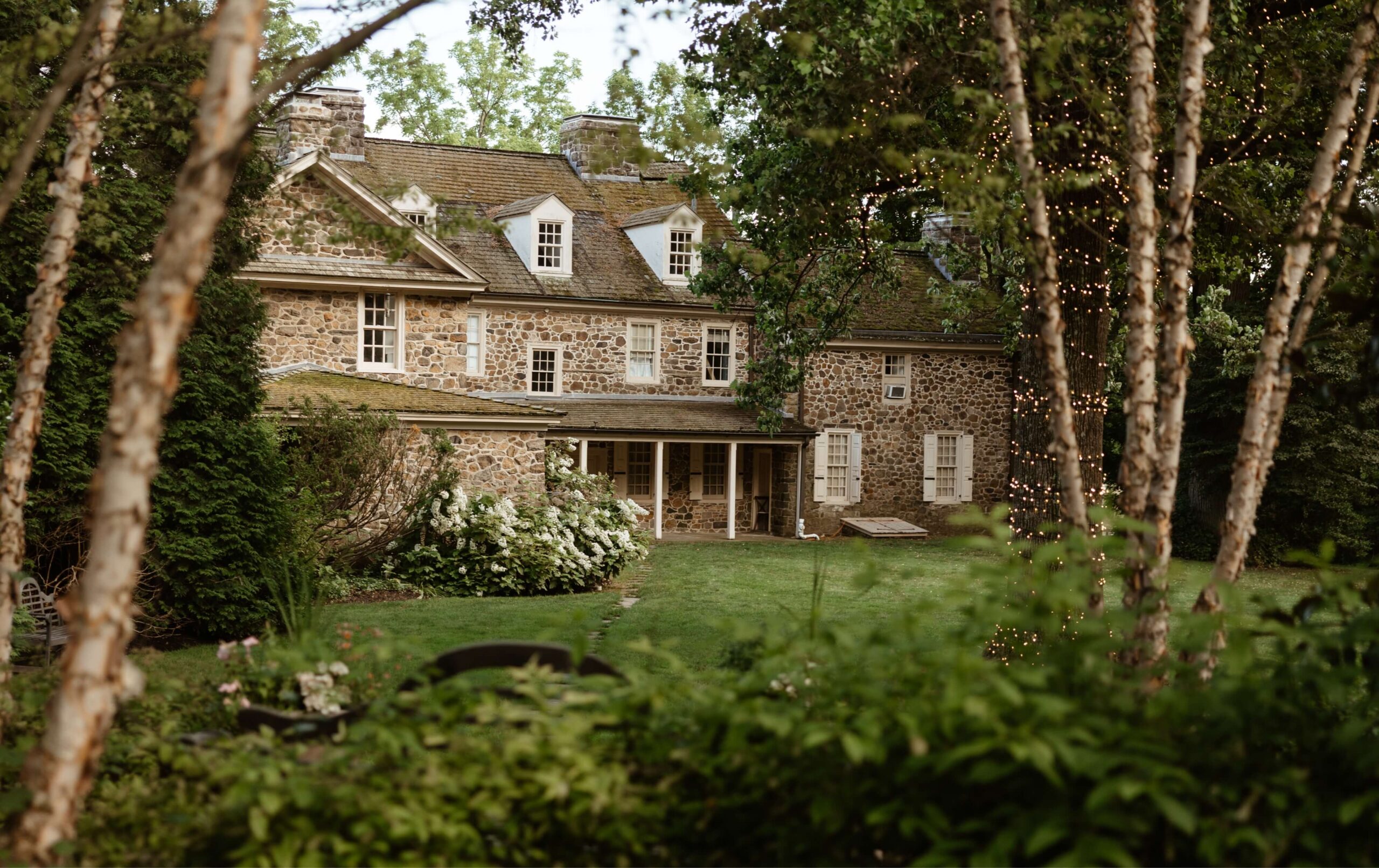 A large stone house with a mossy roof is surrounded by trees and greenery. A patio with pillars is visible, and a tree with string lights is in the foreground.