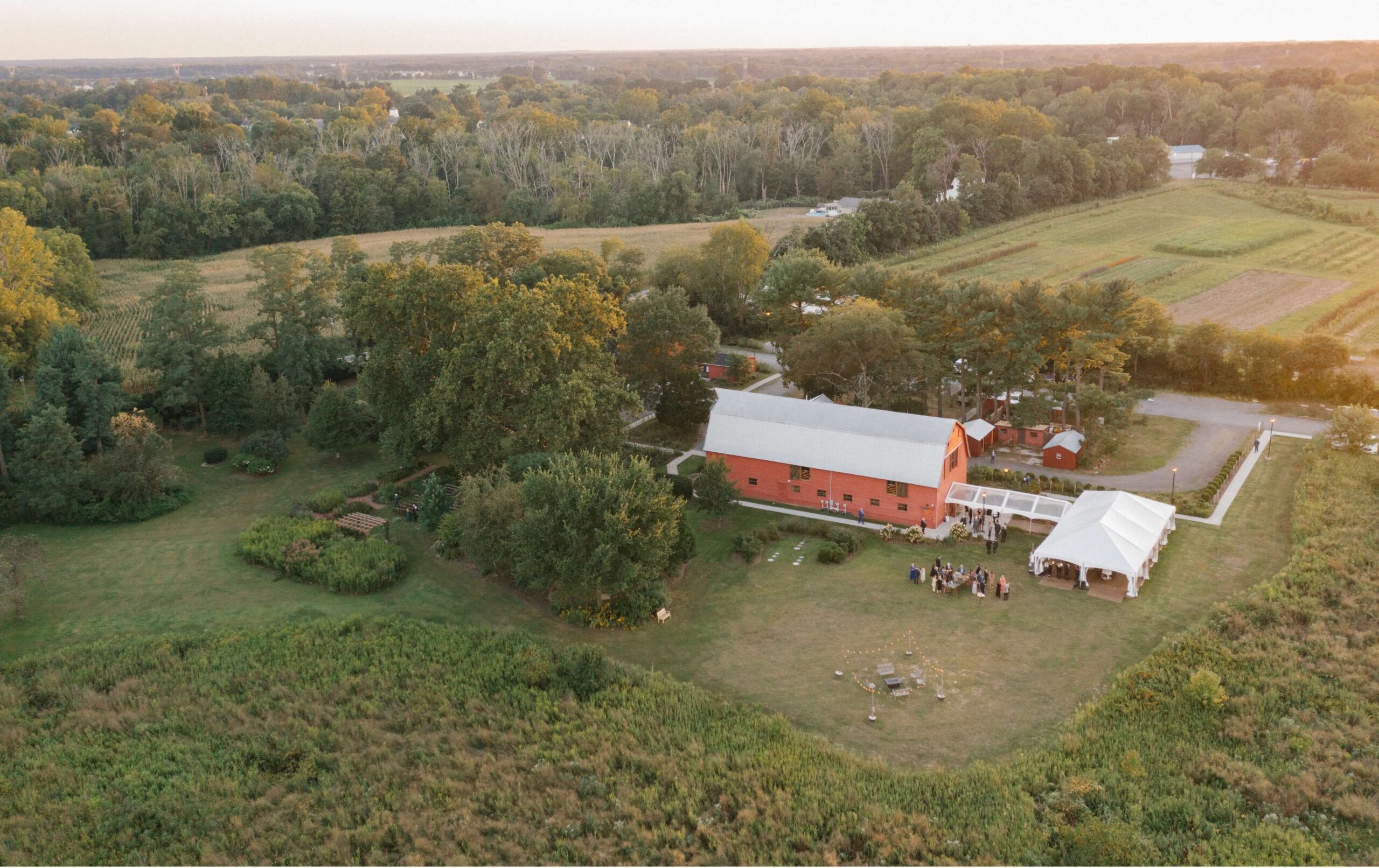 Aerial view of a rural landscape featuring a large red barn, a group gathered under a white tent, surrounded by fields, trees, and a road in the distance, illuminated by sunset light.