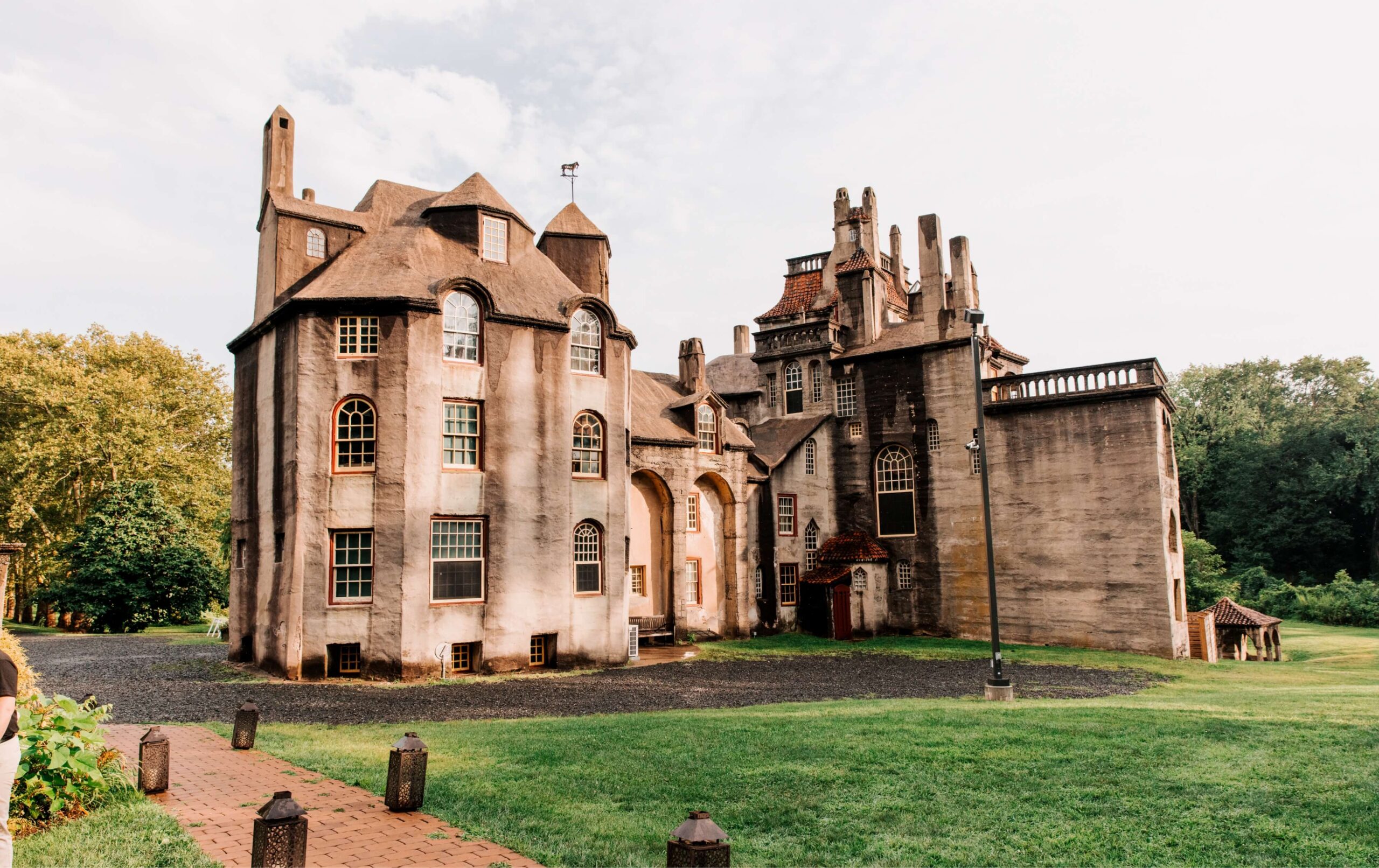 A large, historic stone building with multiple towers and windows, surrounded by a grassy area and trees, under a partly cloudy sky.