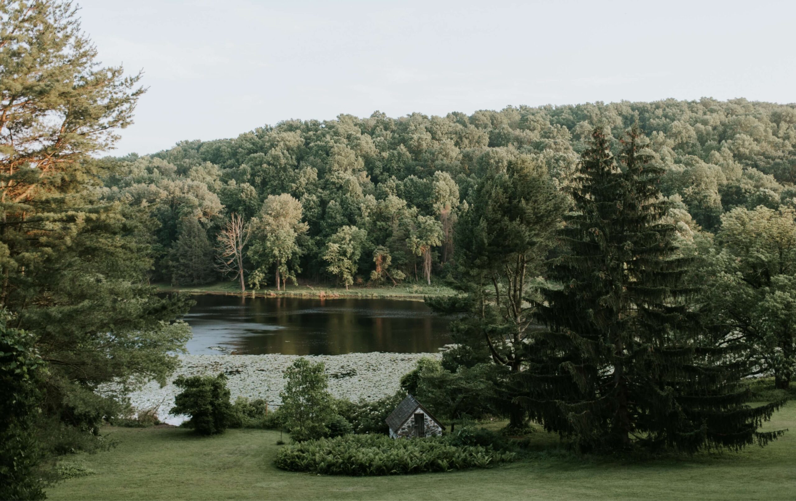 A small stone structure is surrounded by trees and greenery in front of a lake, with a forested hillside in the background under a clear sky.