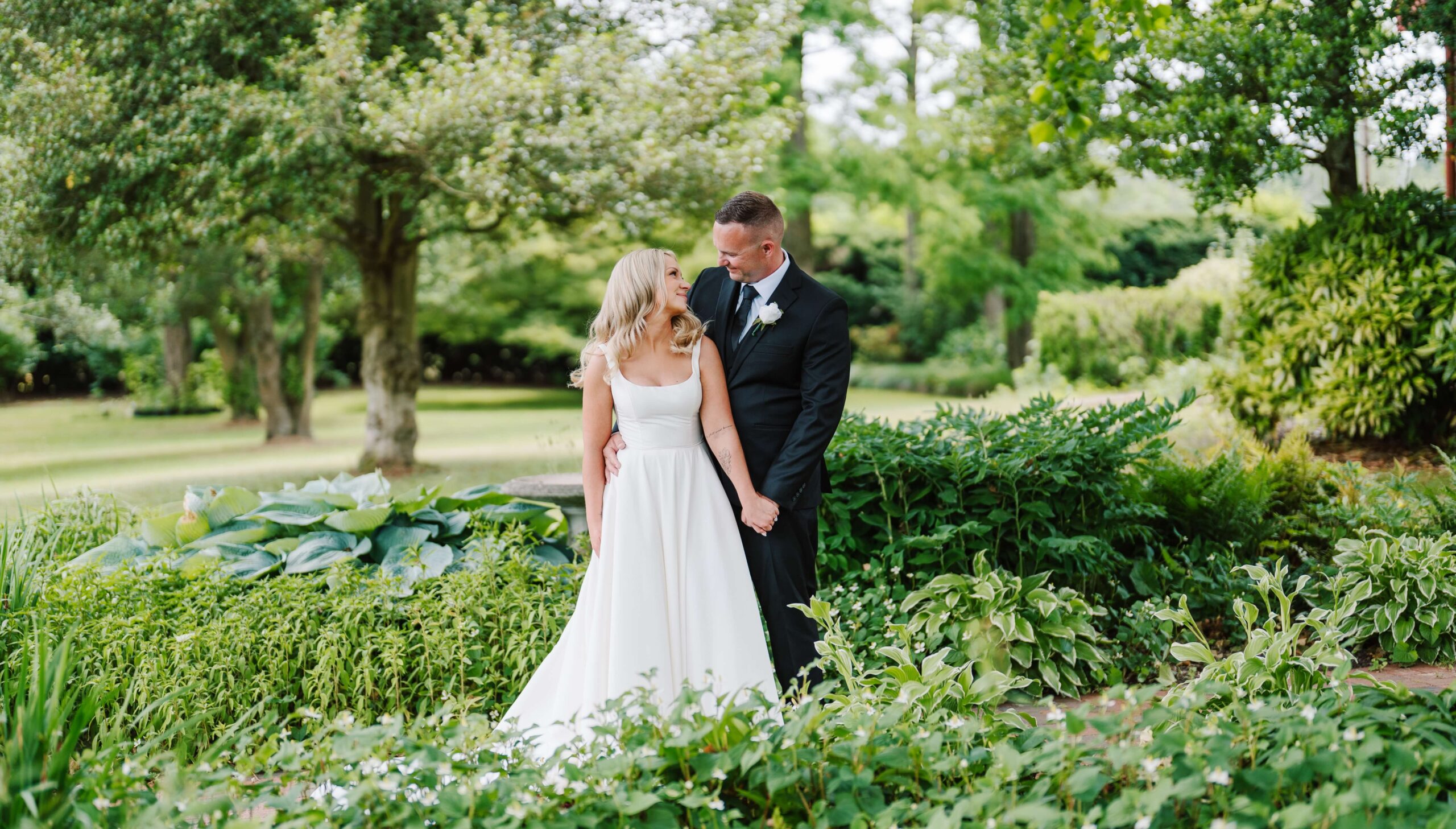 Bride in white dress and groom in black suit stand holding hands in a lush green garden.