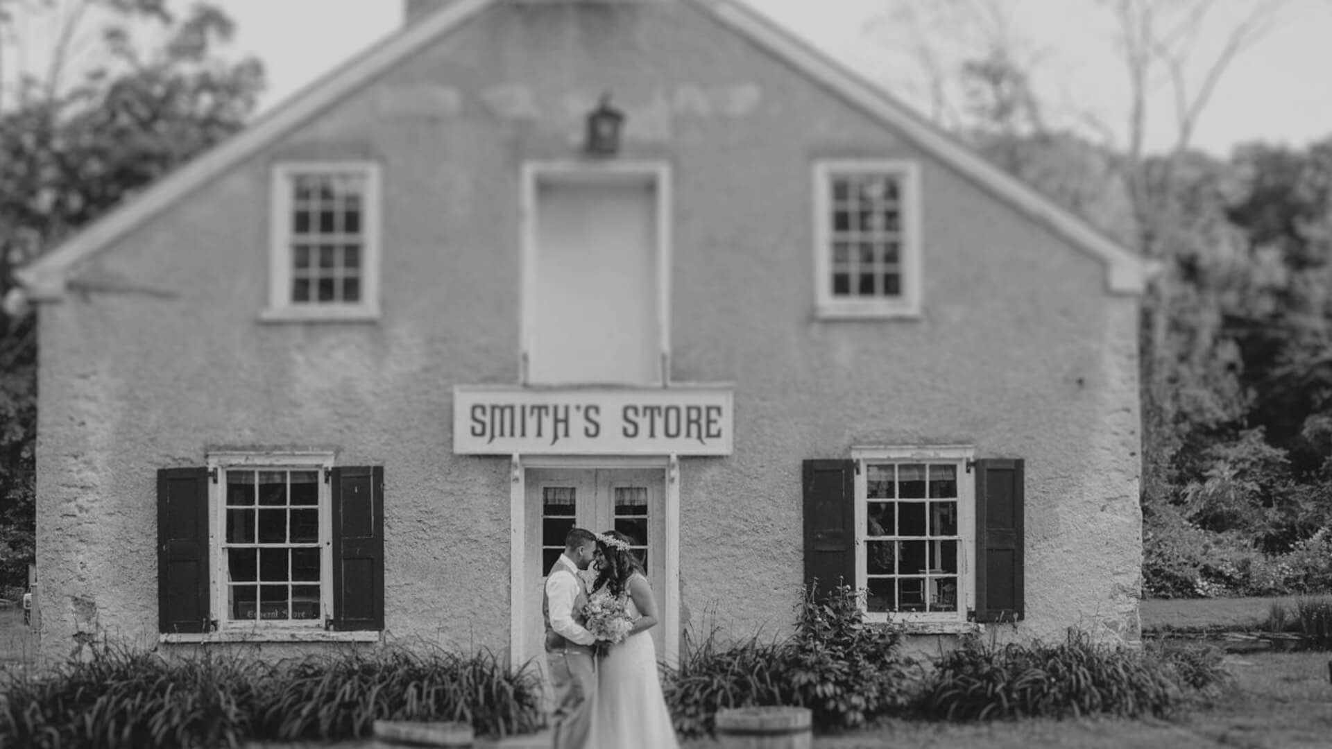 A couple shares a moment in front of a historic building labeled "Smith's Store," surrounded by greenery. The image is in black and white.