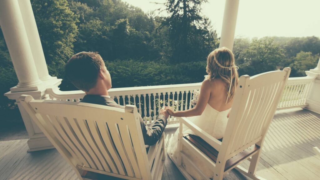A couple sits on wooden rocking chairs holding hands on a porch overlooking lush trees.