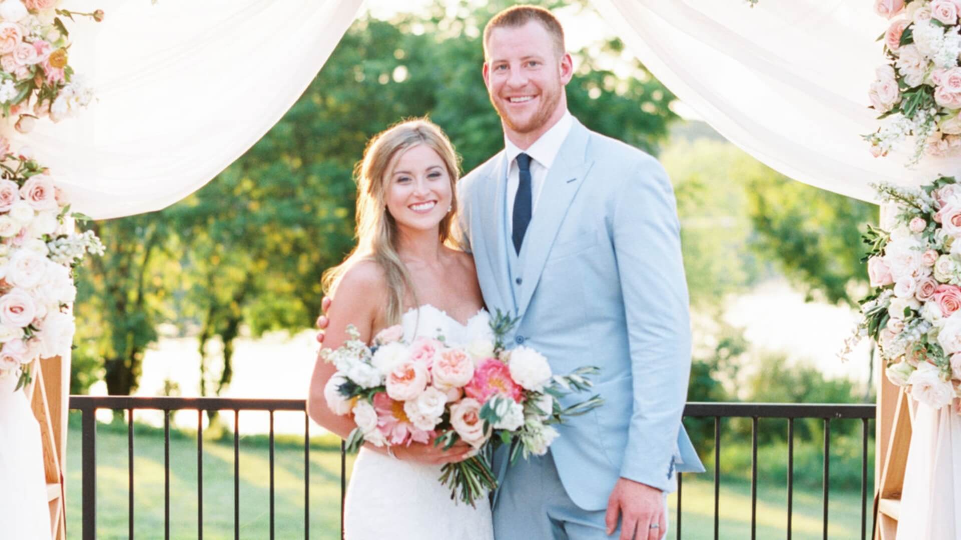 Bride and groom standing under a floral arch, smiling. The bride holds a bouquet of flowers, and the groom wears a light blue suit. Trees are visible in the background.