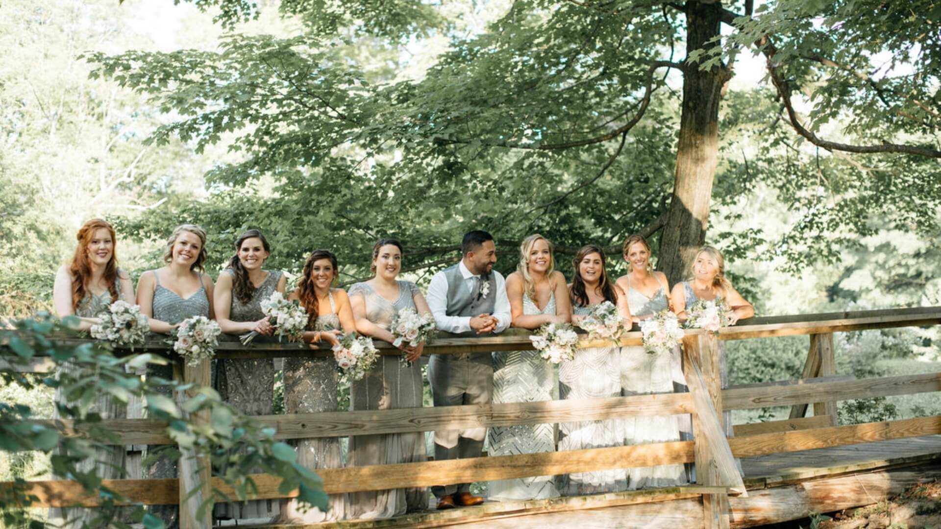 A group of people in formal attire stand on a wooden bridge, surrounded by greenery, holding bouquets.