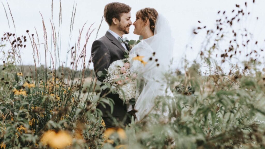 A bride and groom stand smiling at each other in an outdoor setting, surrounded by wildflowers and tall grass.